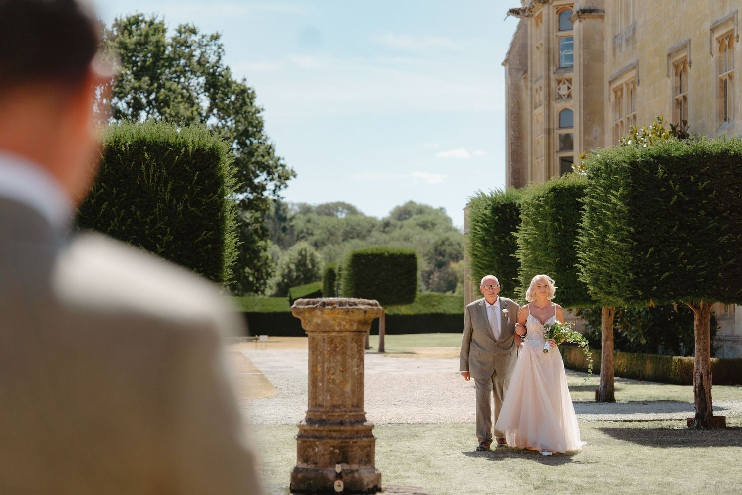 Bride in pink gown walks with escort toward groom at elegant garden wedding venue with manicured hedges.
