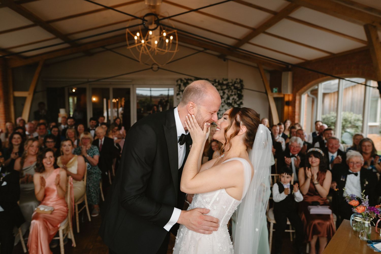 Bride and groom share first kiss at wedding ceremony with guests applauding in elegant venue.
