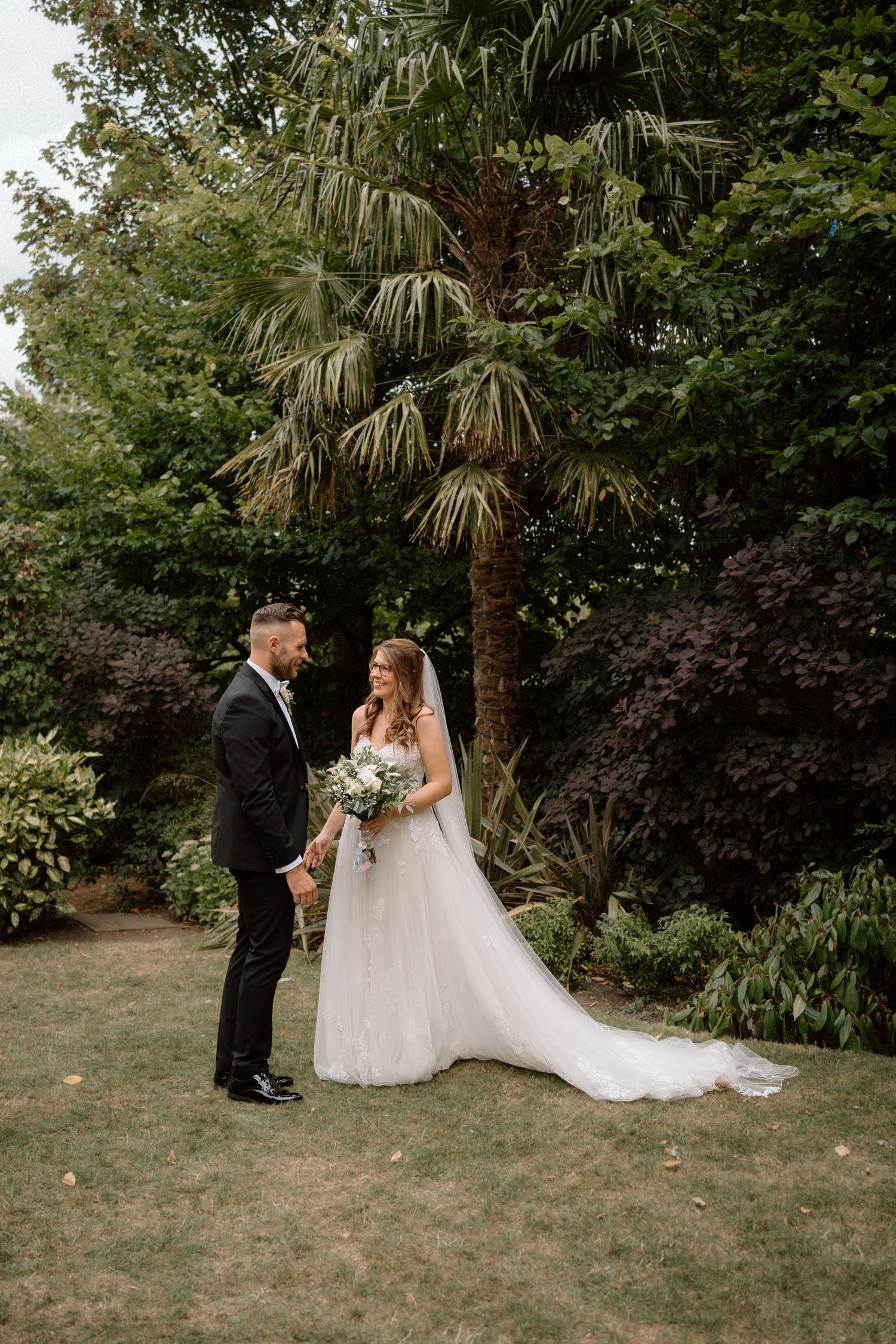 A couple stands together on a lawn with a palm tree and lush garden backdrop during their outdoor wedding ceremony at Warwick House.