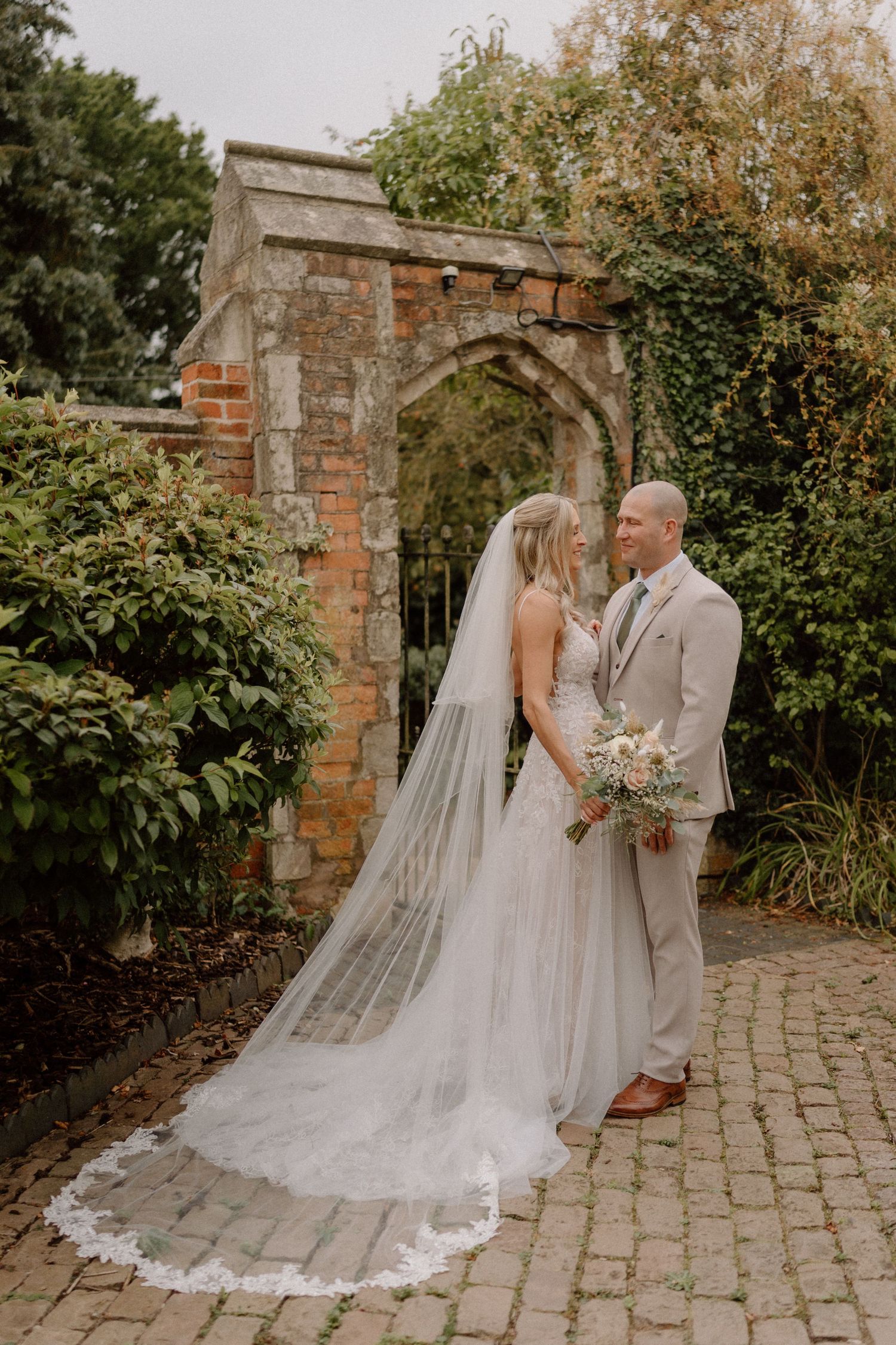A romantic wedding photo showing a couple in front of a rustic stone archway with trailing greenery and flowers in Warwickshire.