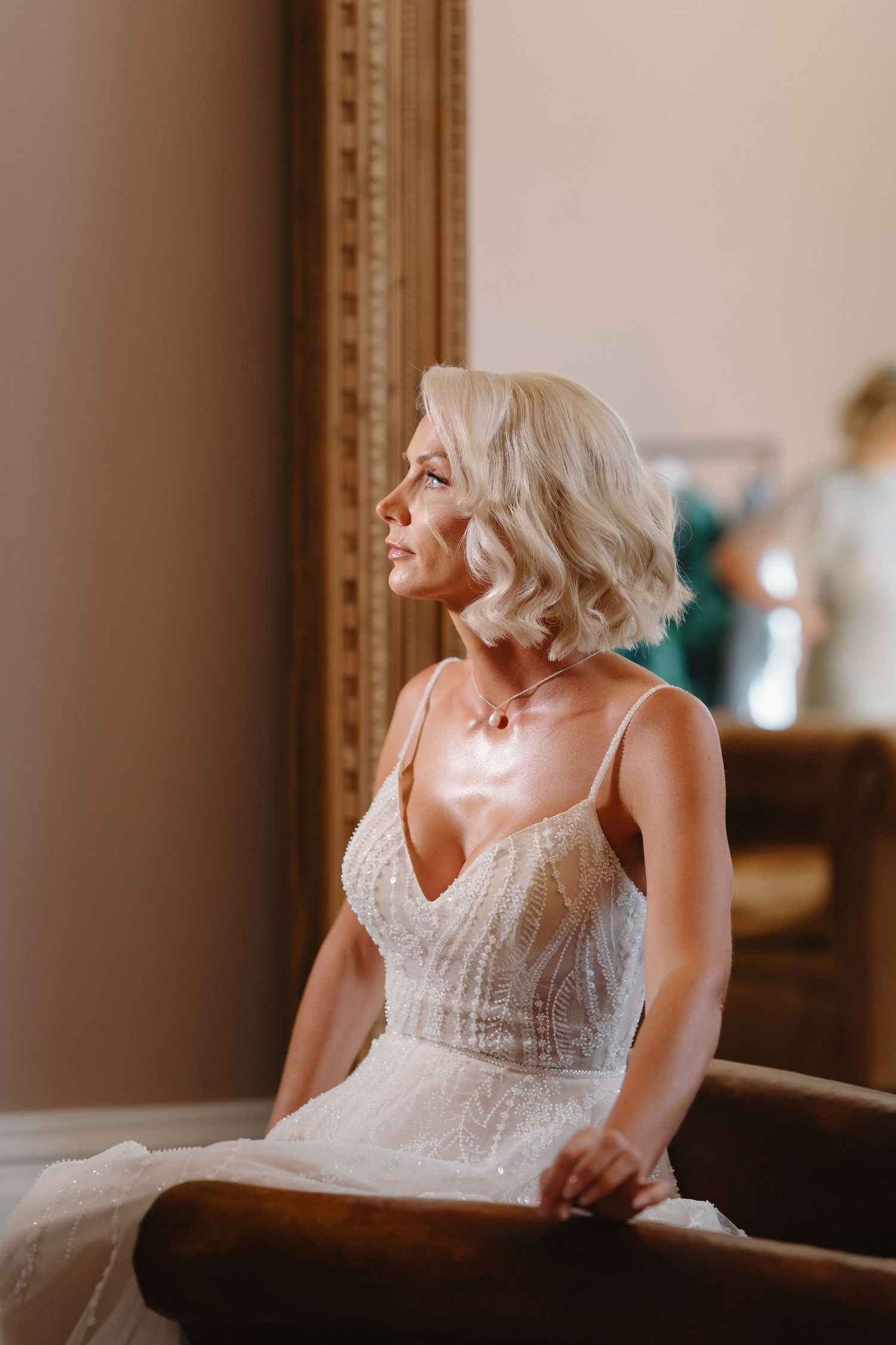 Elegant bride in a lace wedding gown with short blonde hair sitting gracefully near a golden mirror.