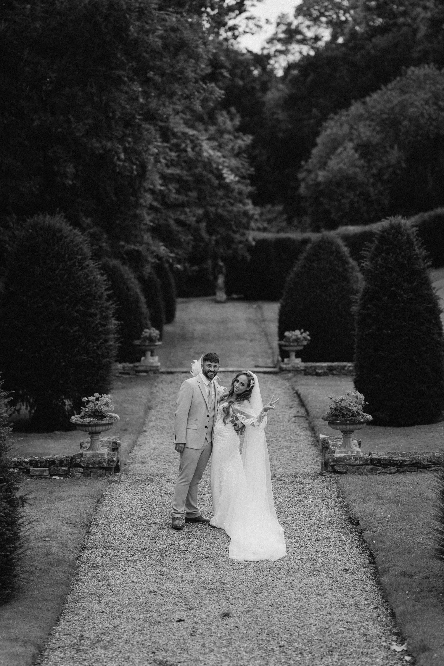 A romantic black and white photo of newlyweds on a formal garden path lined with manicured hedges.