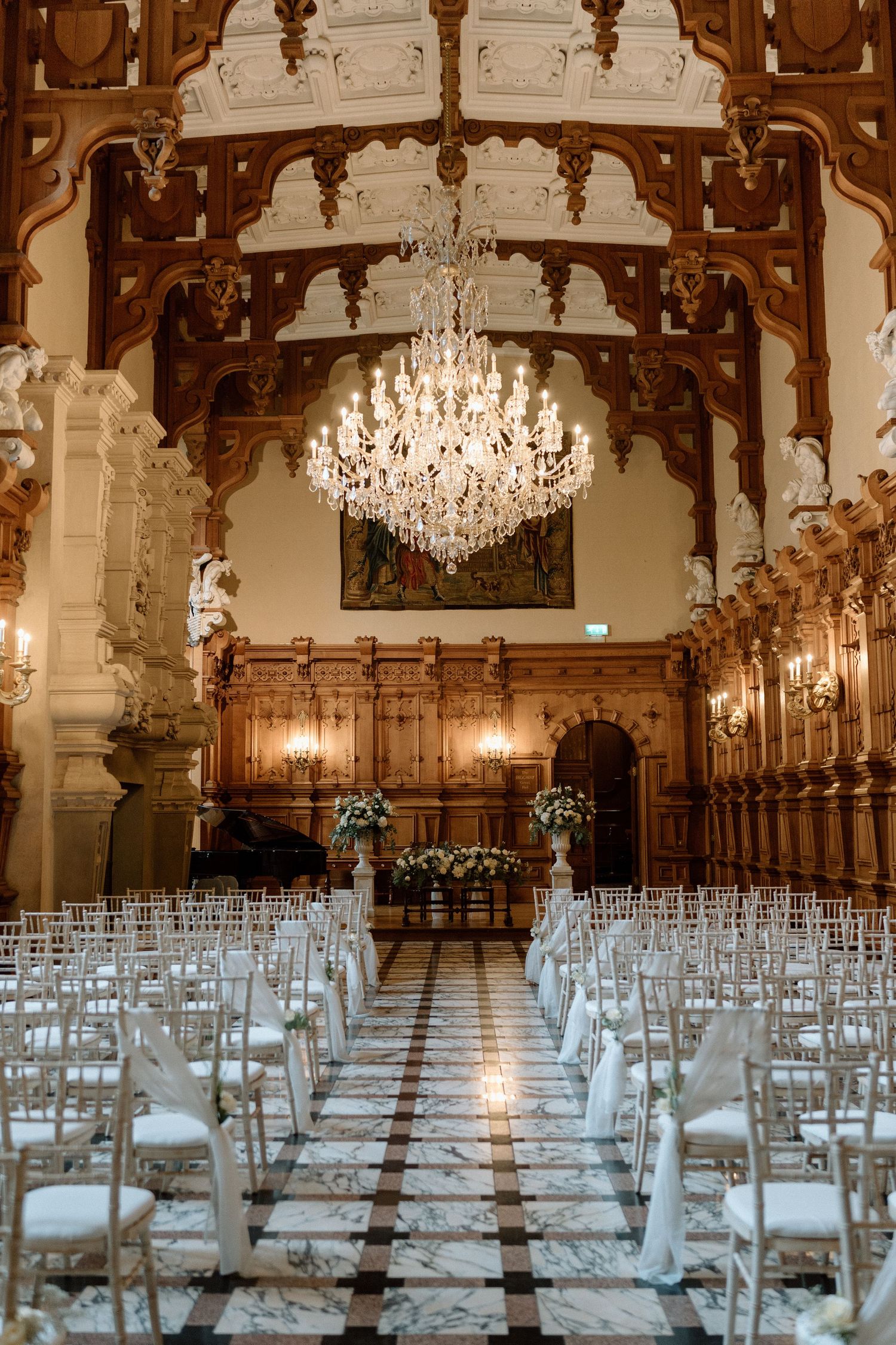 A grand wedding ceremony venue with ornate wooden arches, crystal chandelier and rows of white chairs at Harlaxton Manor.