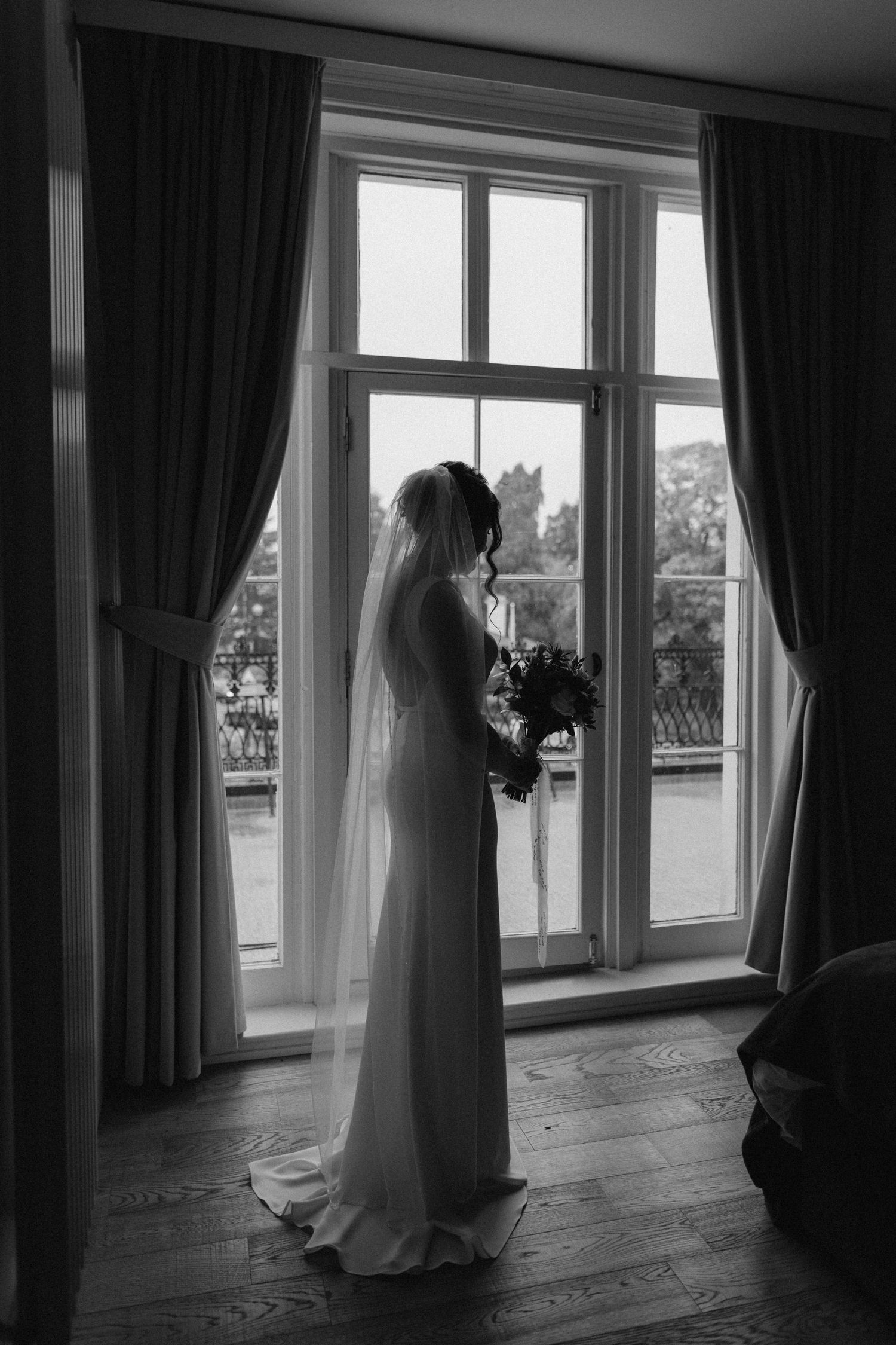 A silhouetted figure in a wedding dress stands by a large window with flowing curtains in Leamington Spa.