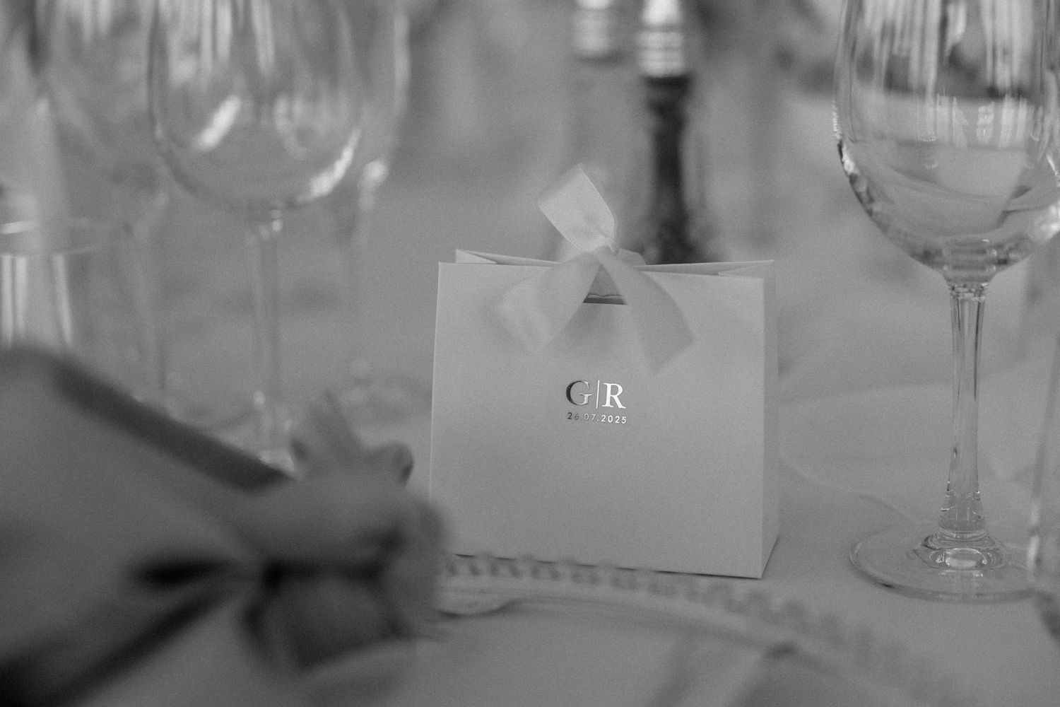 An elegant wedding favor bag with initials CR sits between crystal wine glasses on a formal table setting.
