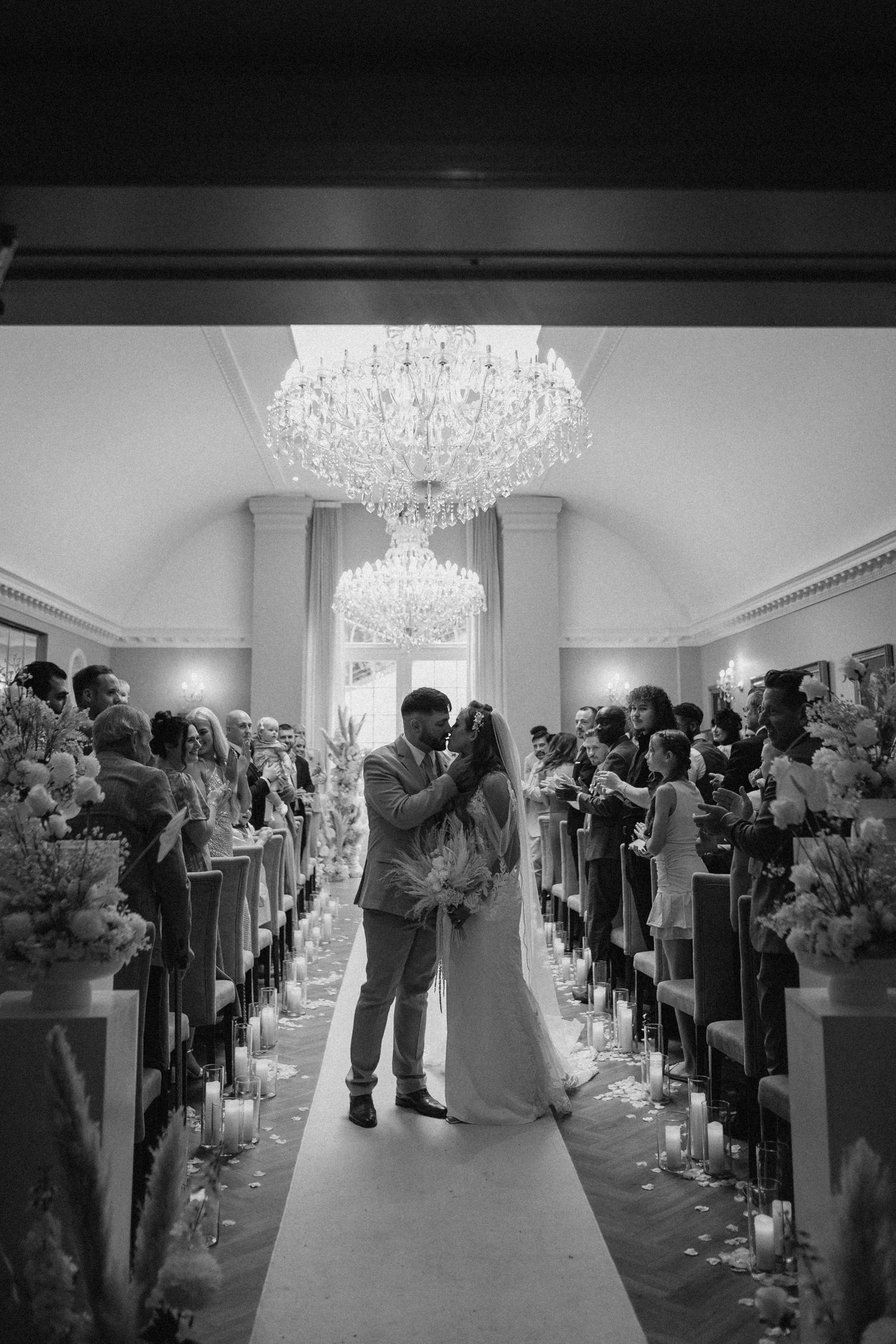 A wedding ceremony moment under crystal chandeliers with candle-lined aisle and floral arrangements.