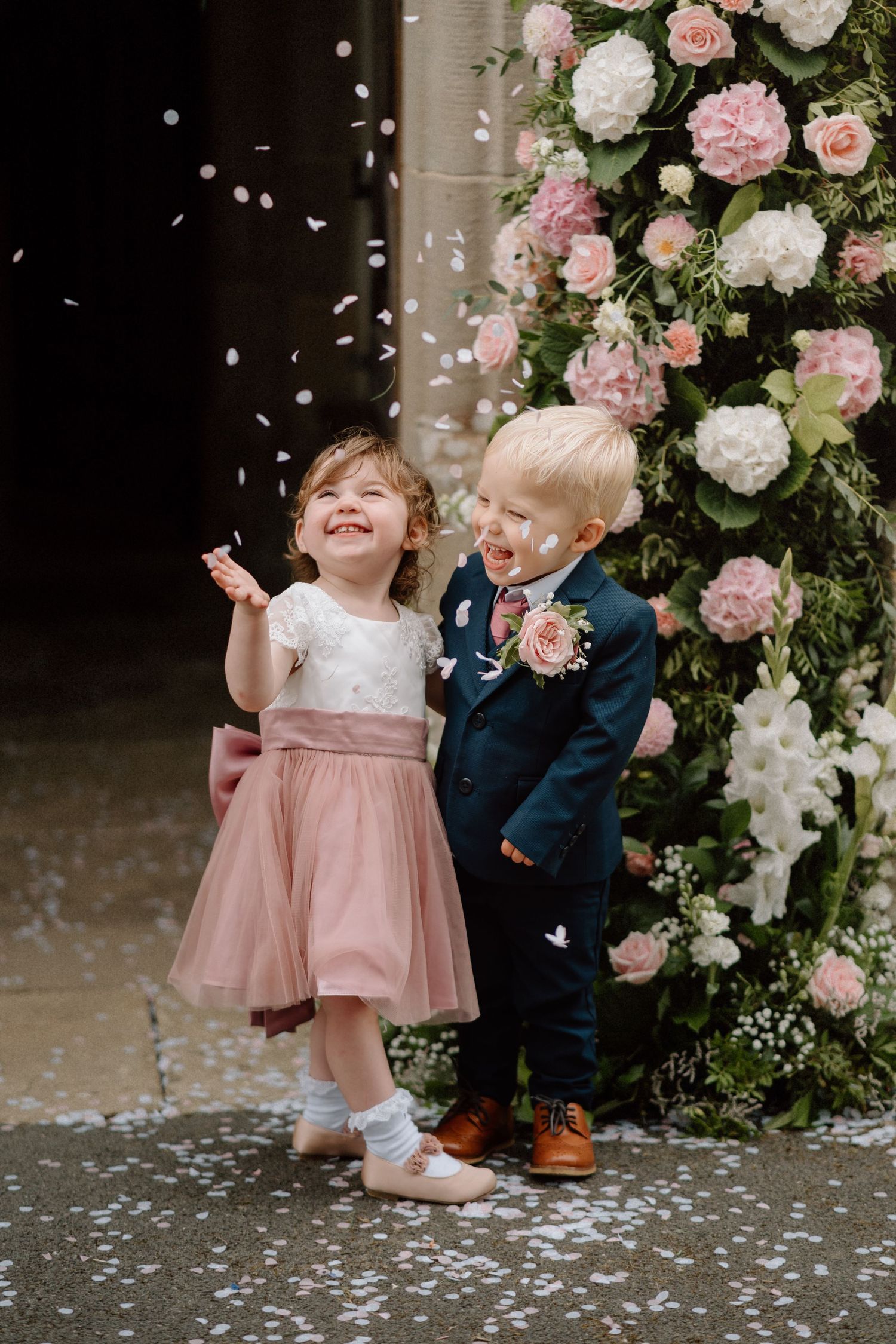 Two young children stand together by pink and white floral arrangement while rose petals fall around them.