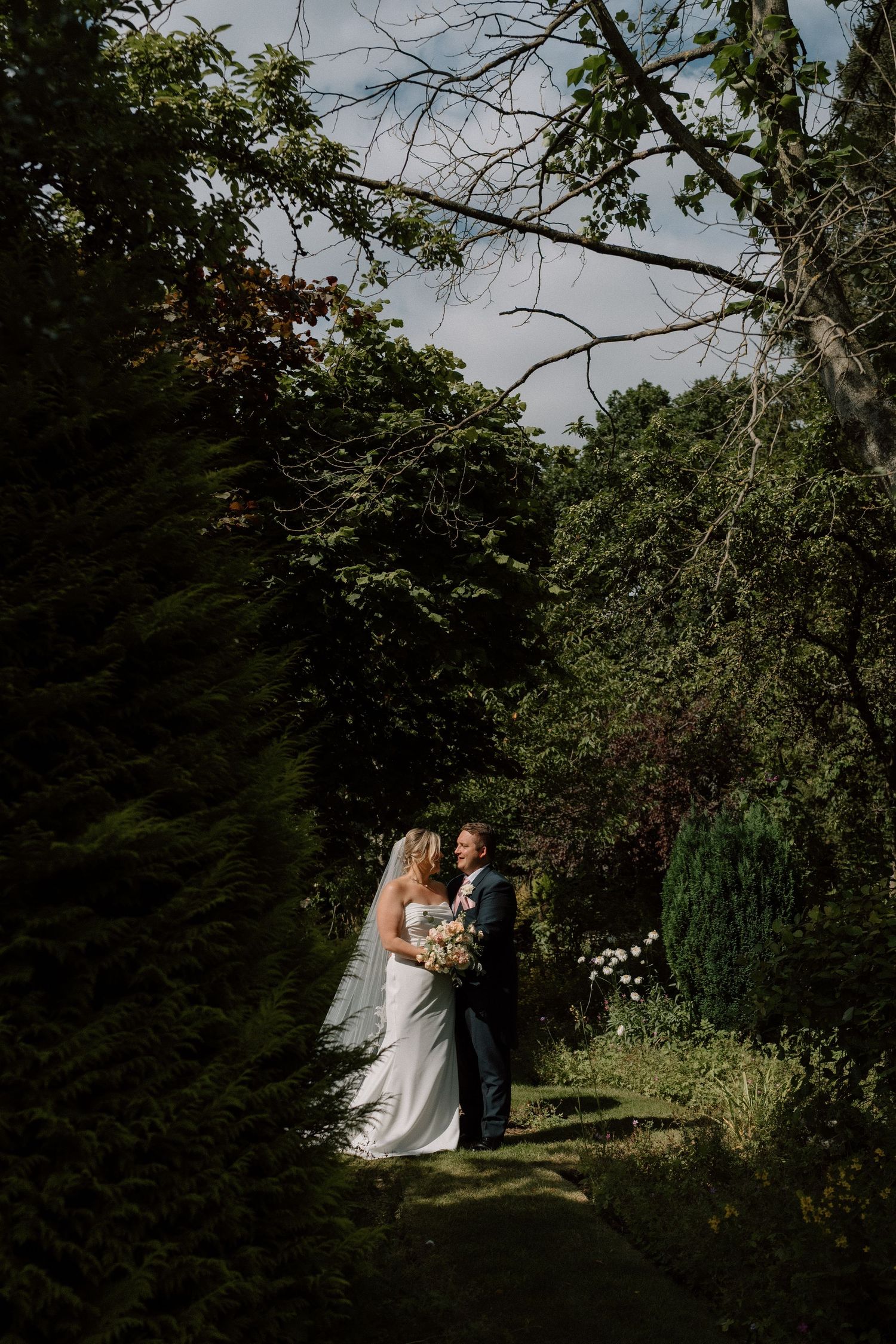 A wedding couple poses on a garden path surrounded by lush green trees and natural scenery at Nuthurst Grange.