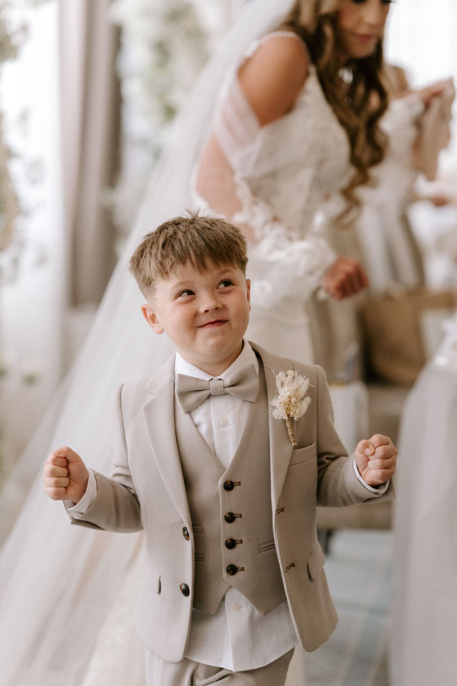 A young boy in a light gray three-piece suit with a bow tie smiles while standing next to flowing white wedding dress fabric.