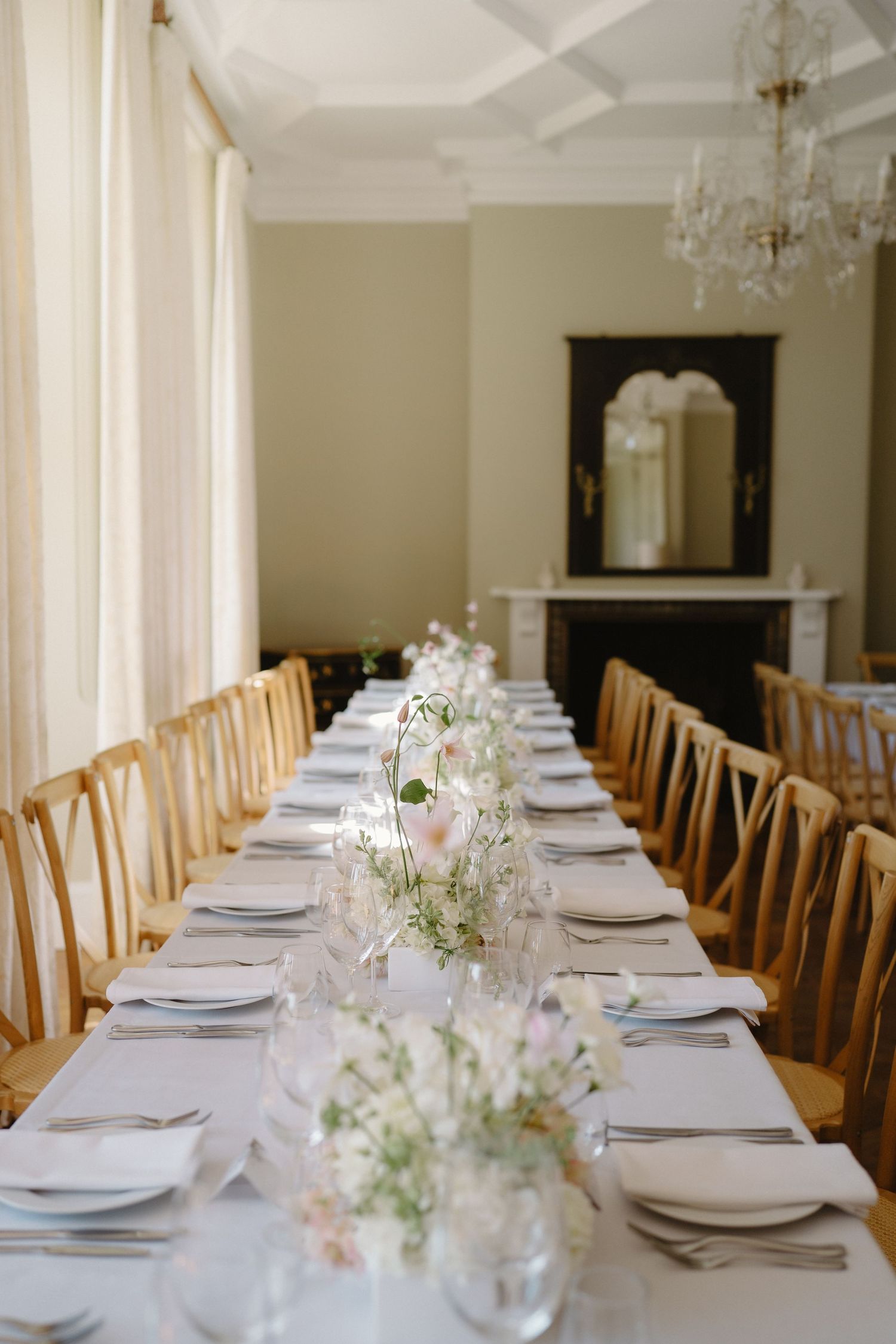Elegant wedding reception table with white linens, floral centerpieces, crystal chandelier, and wooden chairs in a bright dining room.