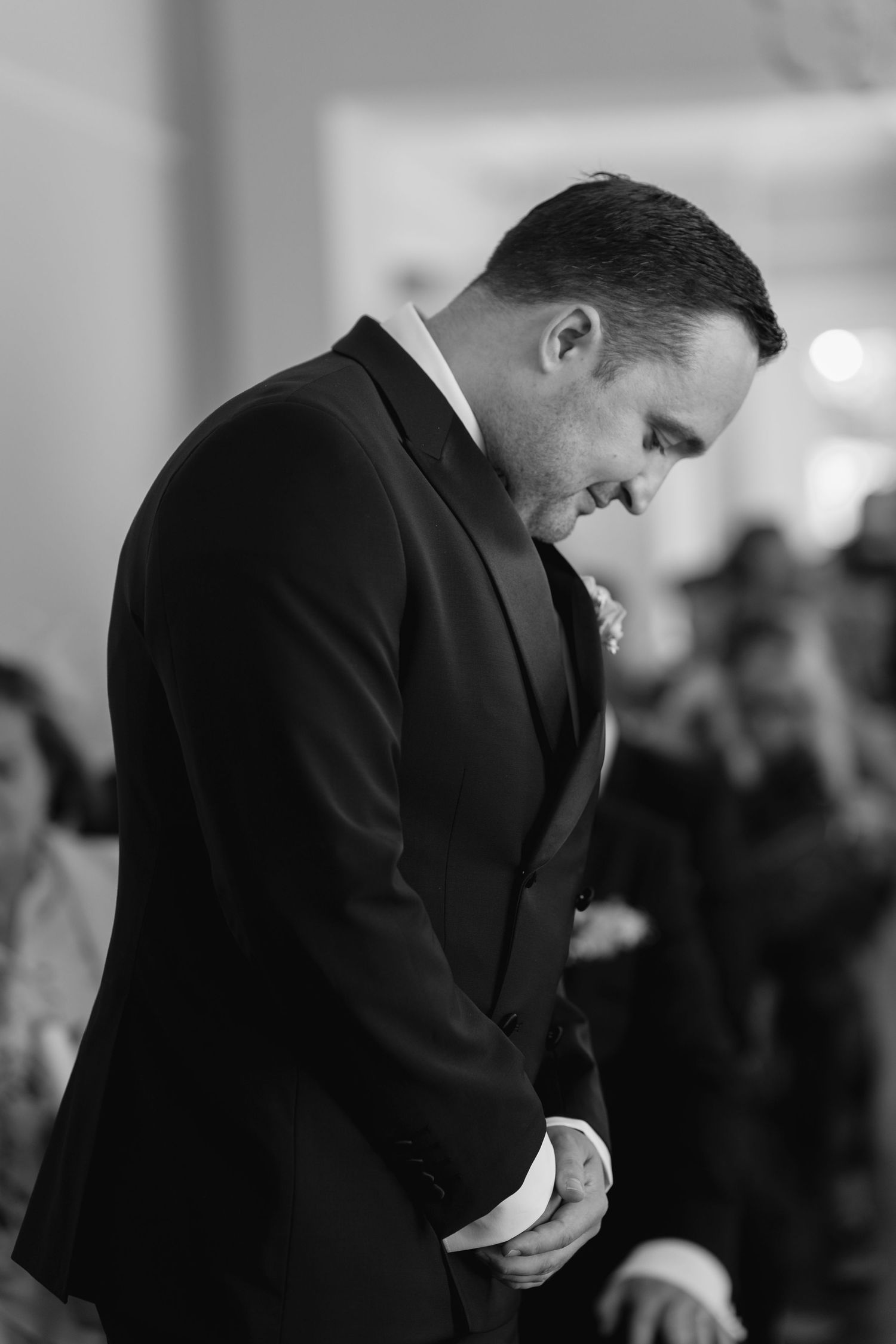 Groom in black tuxedo bowing head solemnly at wedding altar in black and white photo.