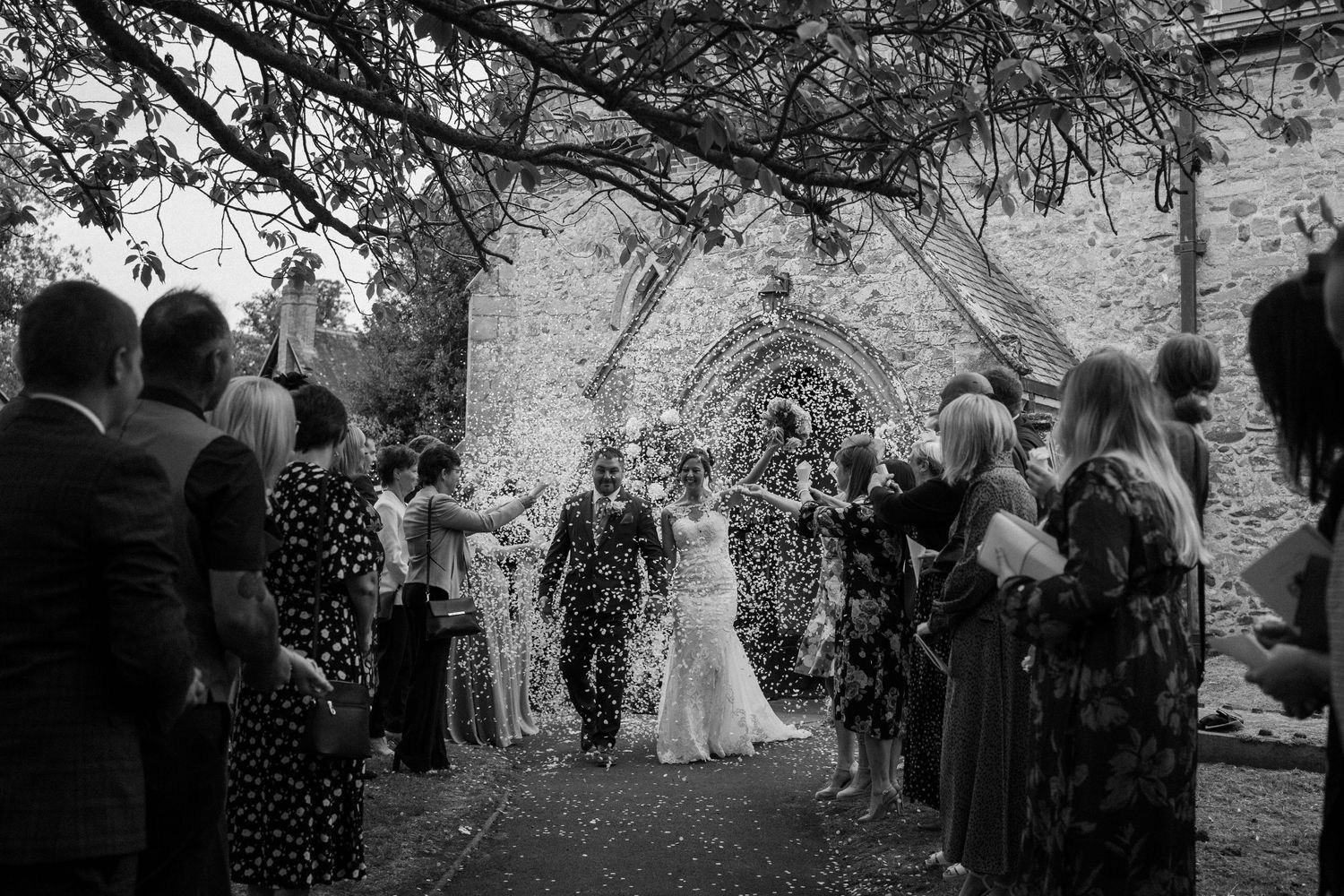 Black and white photo of wedding guests celebrating as the newlyweds walk through a shower of confetti under stone archway.