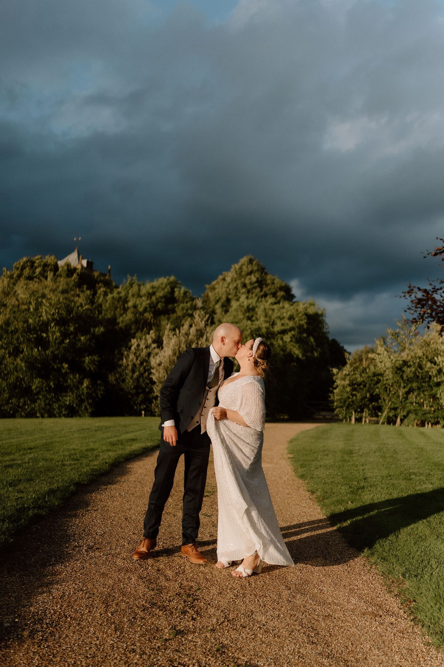 A couple shares an intimate moment on a gravel path under dramatic storm clouds at sunset at Dodford Manor.