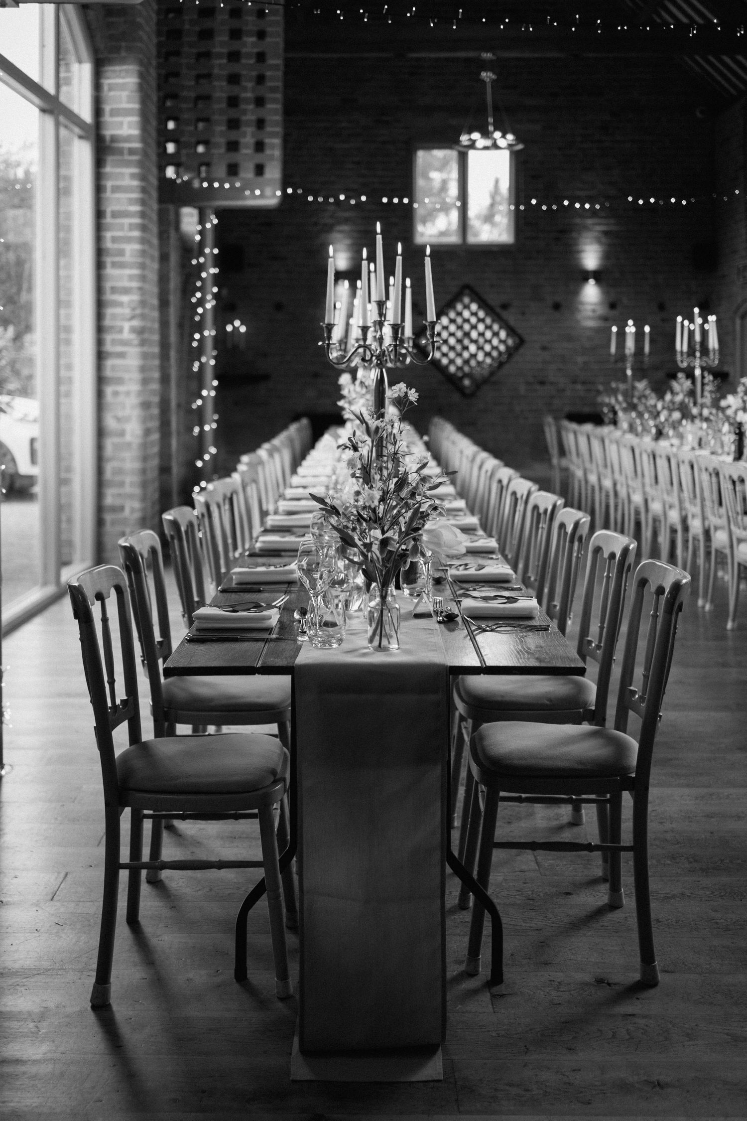 A long elegant dining table set with candelabras and place settings in a rustic brick-walled venue with string lights at Swallows Nest Barn.