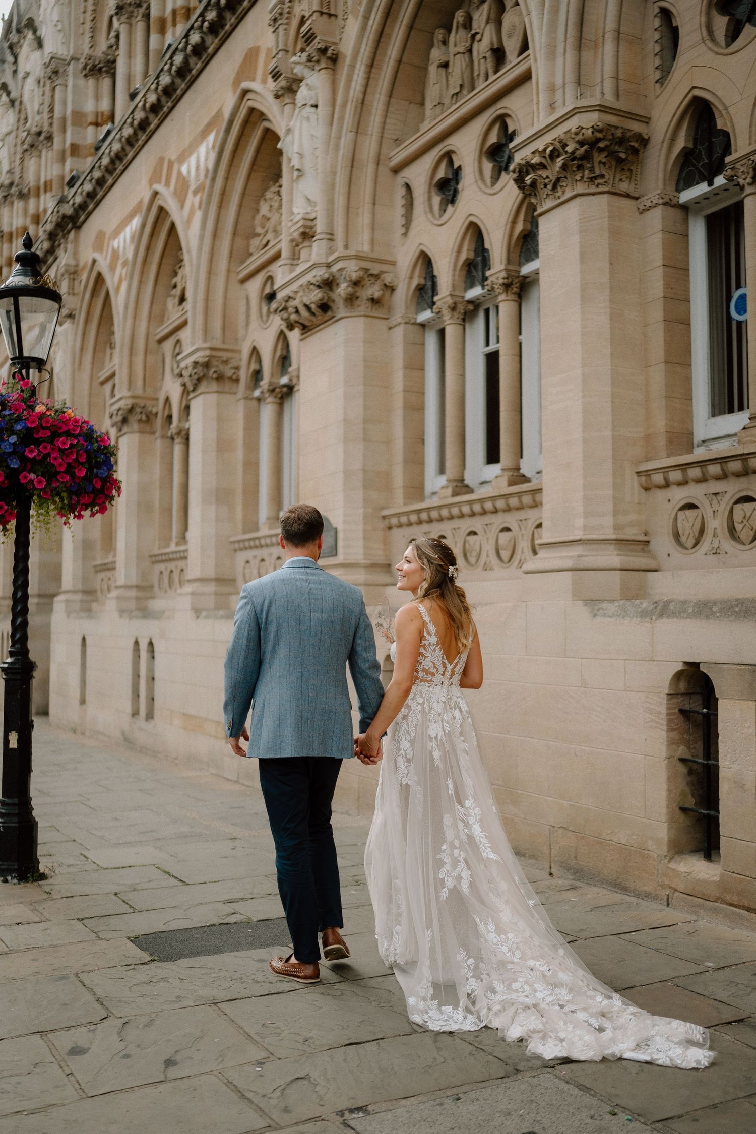 A couple walks hand in hand along a gothic architecture building with trailing wedding dress.