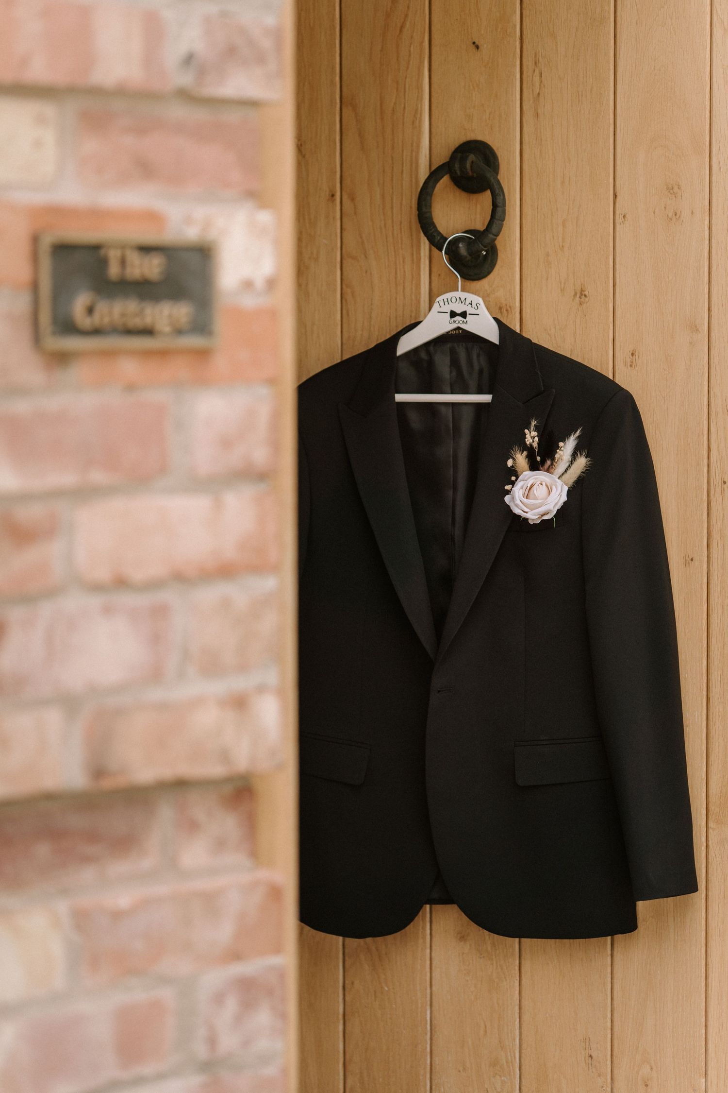 Black formal suit jacket with delicate pink floral boutonniere hangs against wooden paneled wall.
