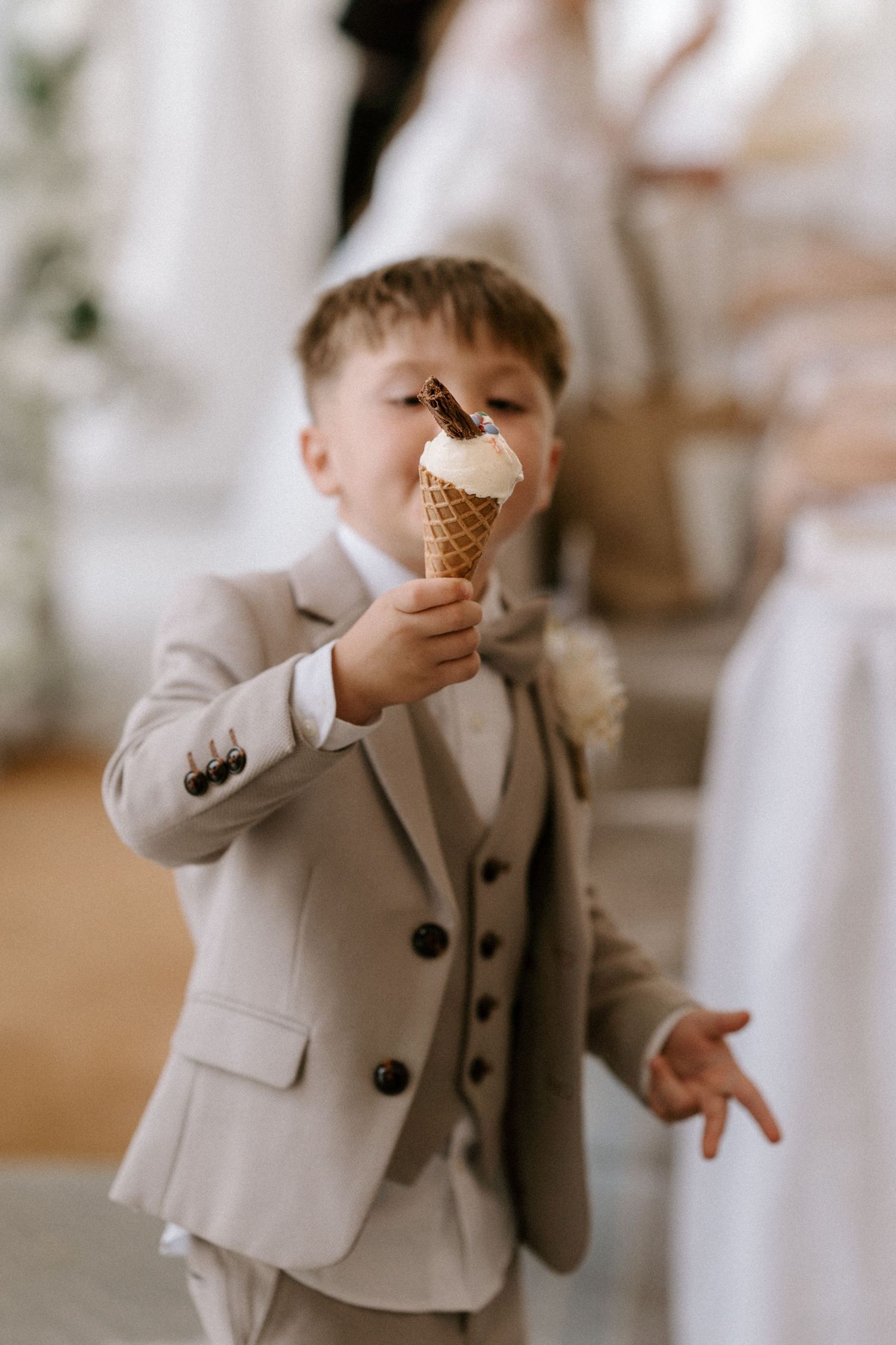 A young ring bearer in a light gray suit enjoying an ice cream cone at a wedding reception.