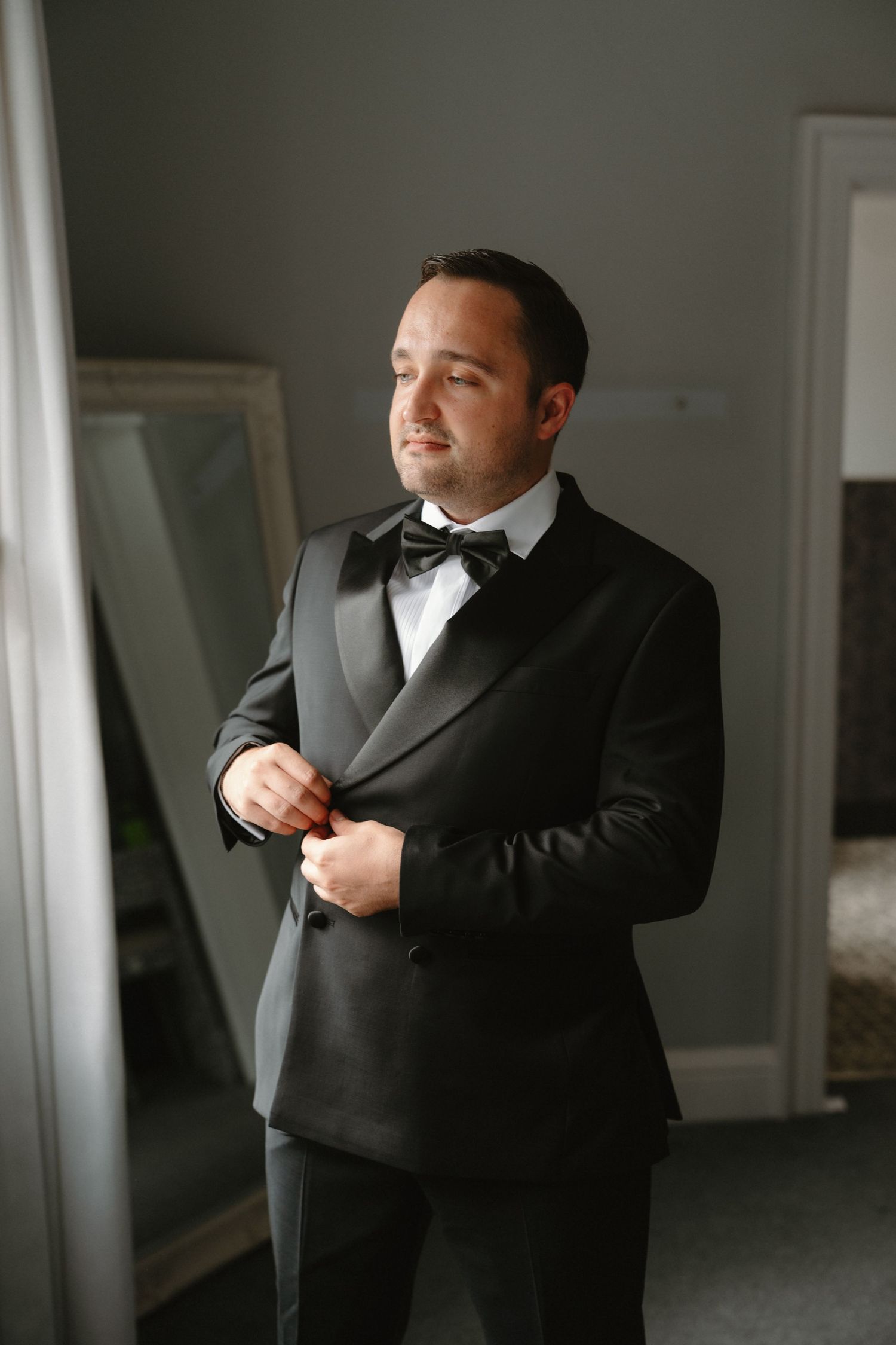 Groom in black tuxedo with bow tie buttoning jacket while standing near mirror before wedding ceremony.
