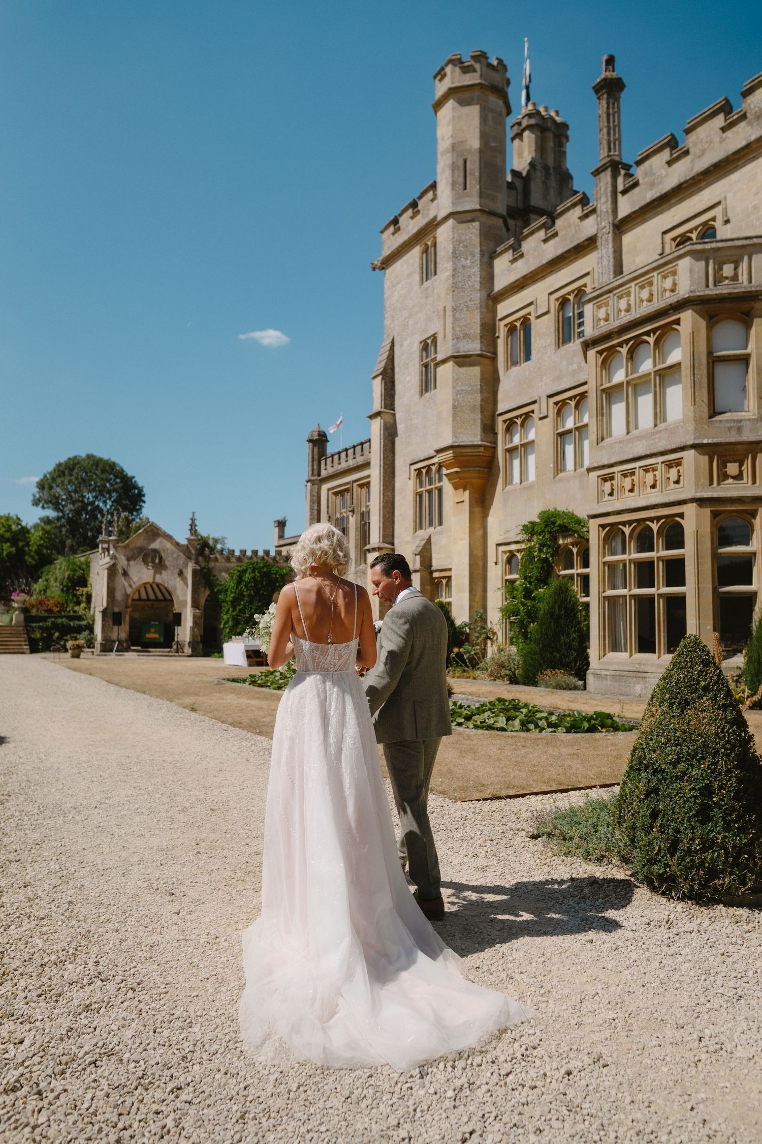 Bride and groom stand before a grand English manor house on a sunny wedding day, surrounded by manicured gardens.