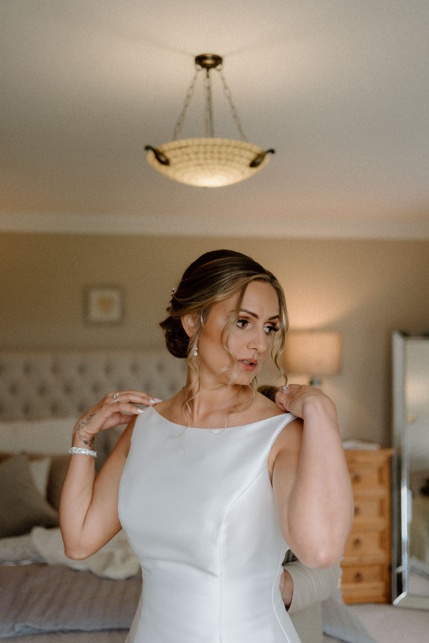 Elegant sleeveless white wedding gown shown in a bedroom with soft lighting and a decorative ceiling light fixture at Dodford Grange