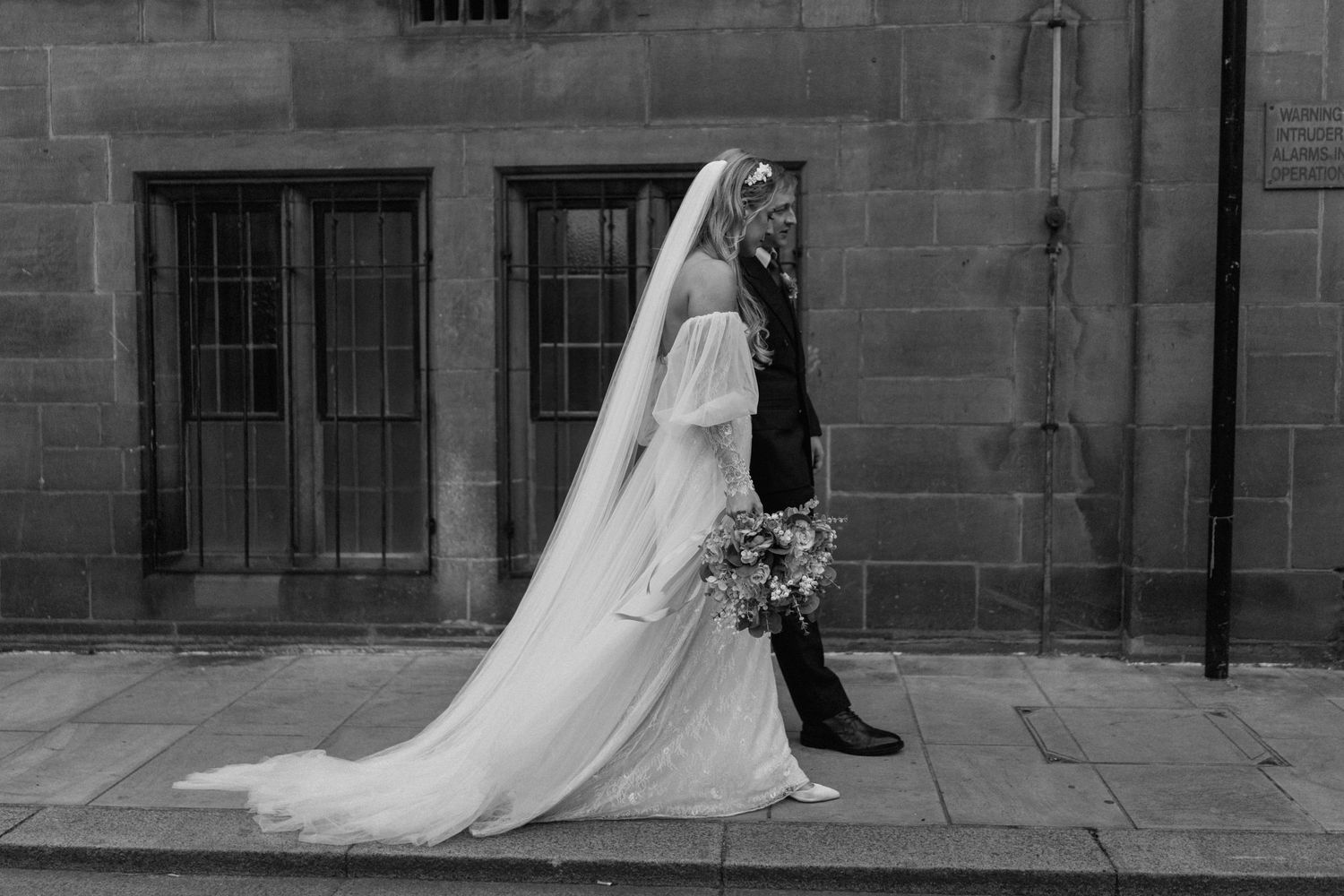 Elegant bride in flowing white gown and veil poses against historic stone church architecture.
