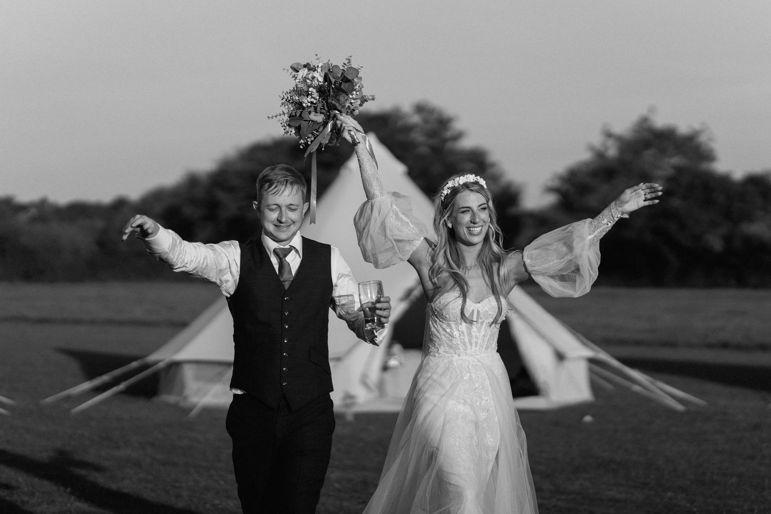 A joyful couple celebrates with arms outstretched in a field at sunset.