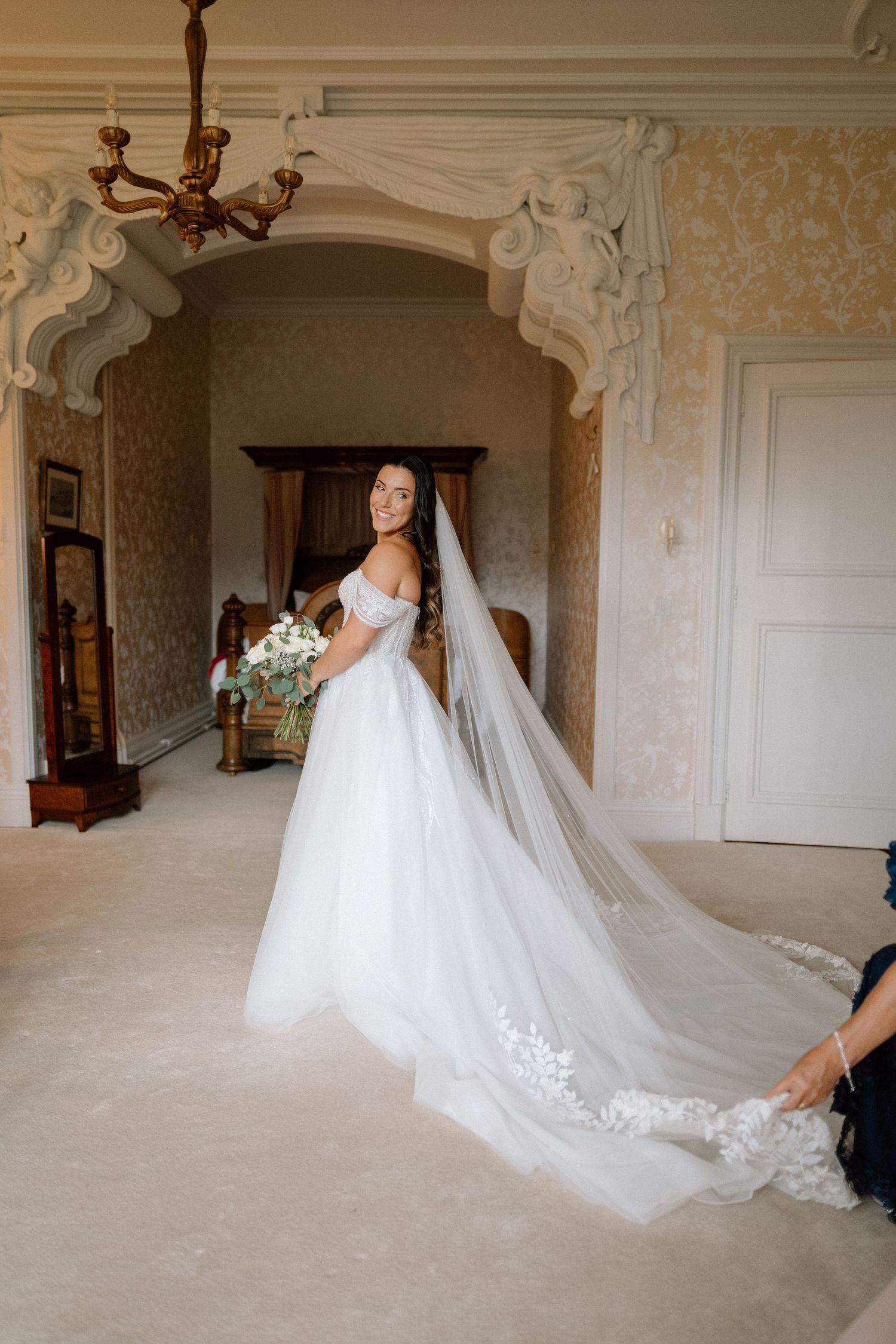 An elegant bridal portrait in a classic interior with detailed wallpaper and flowing cathedral veil at Harlaxton Manor.