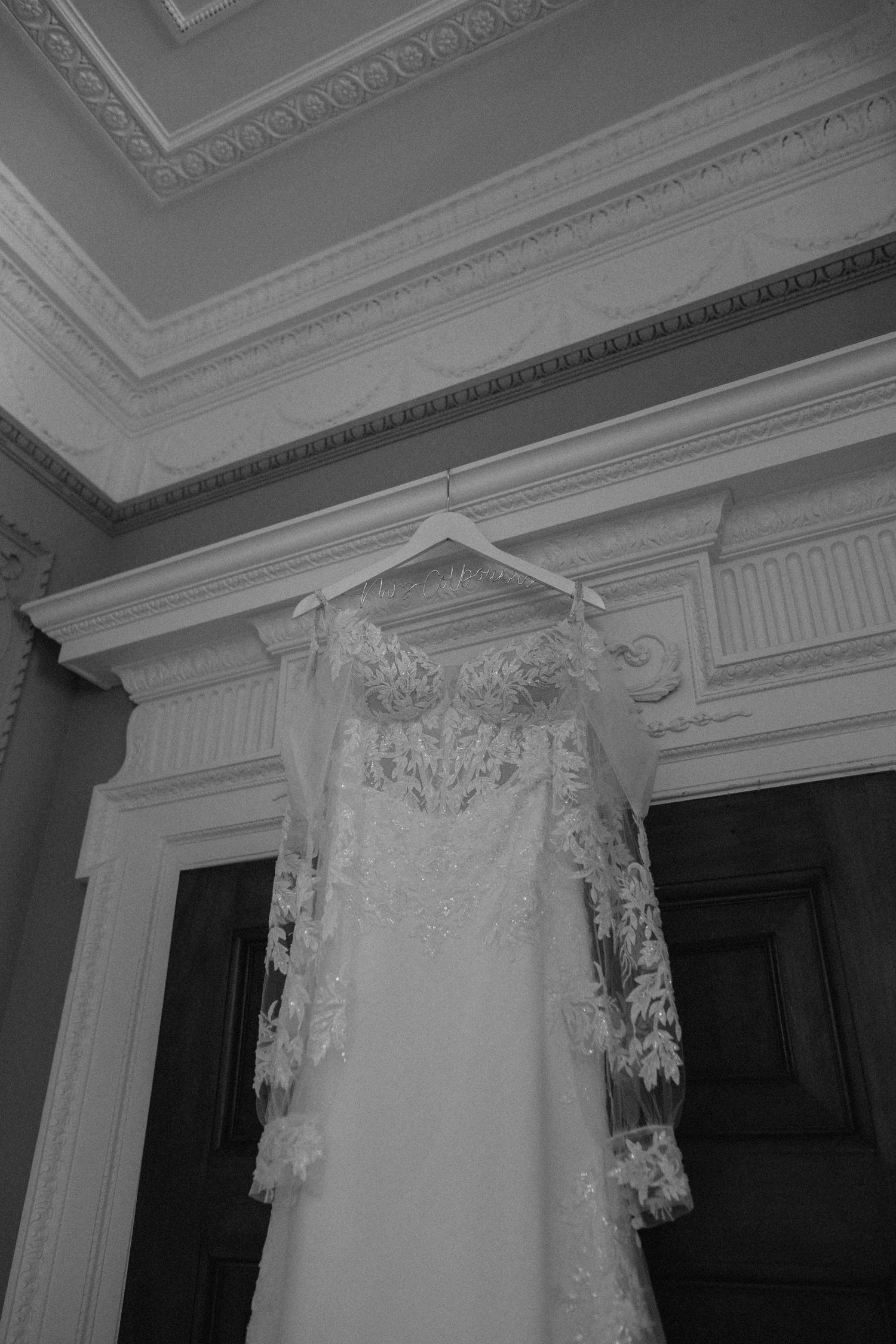 Elegant white lace wedding dress with intricate details hanging against ornate crown molding.