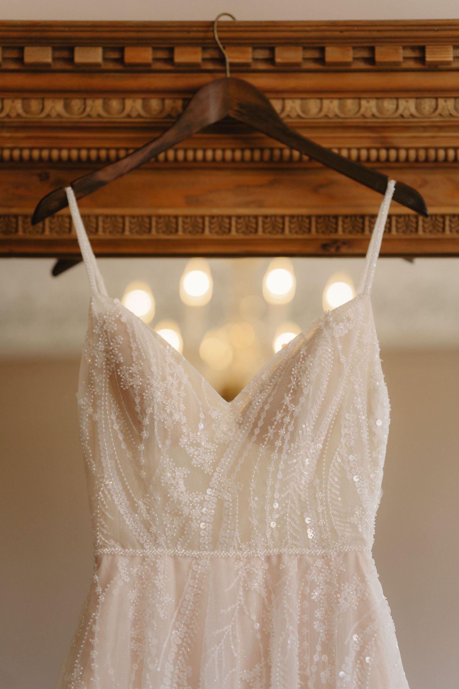 Beaded ivory wedding dress with spaghetti straps on wooden hanger against ornate gold mirror.