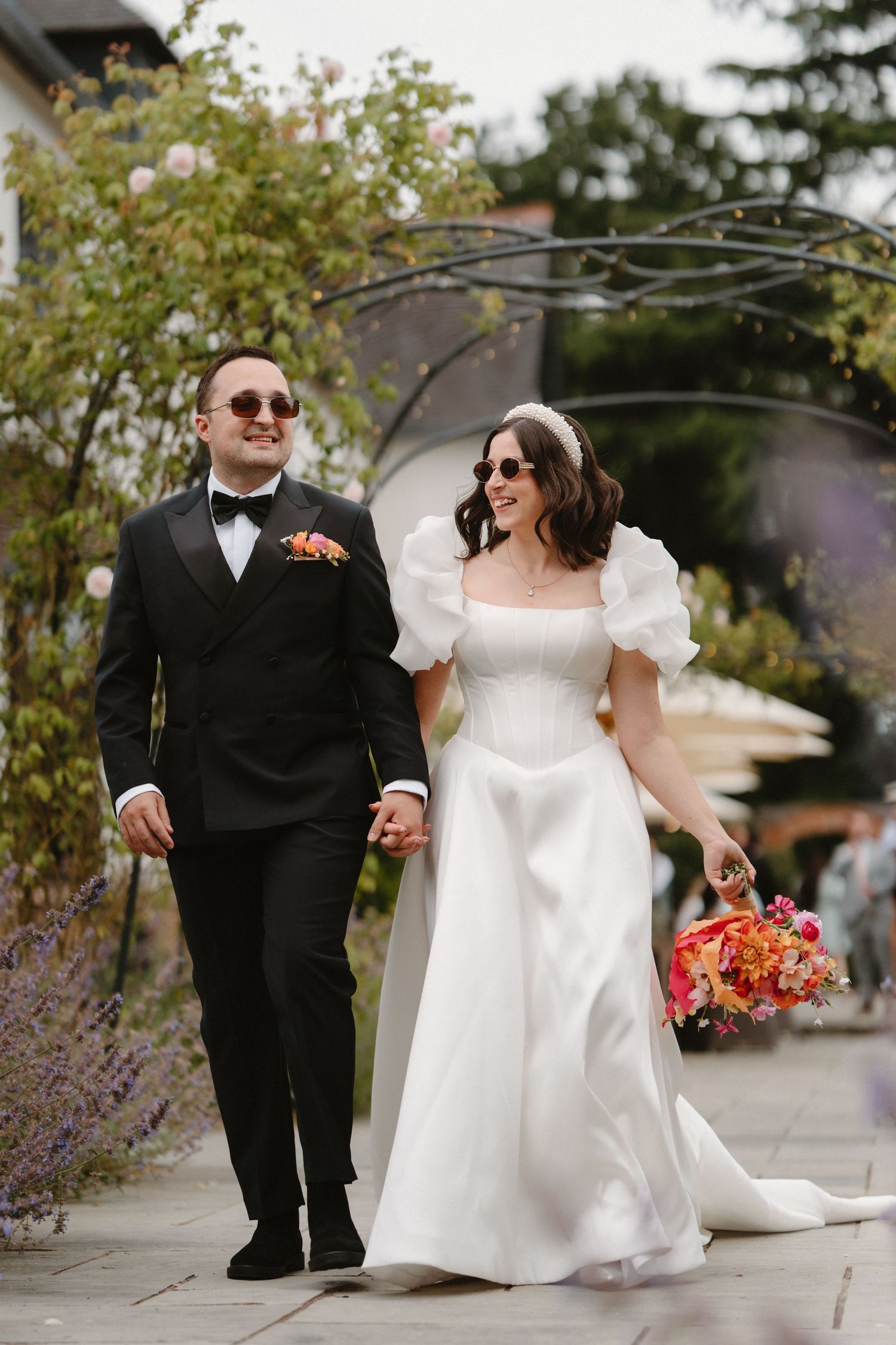 Bride and groom walk hand-in-hand in a garden, she in a white puff-sleeve gown, he in a black tuxedo.