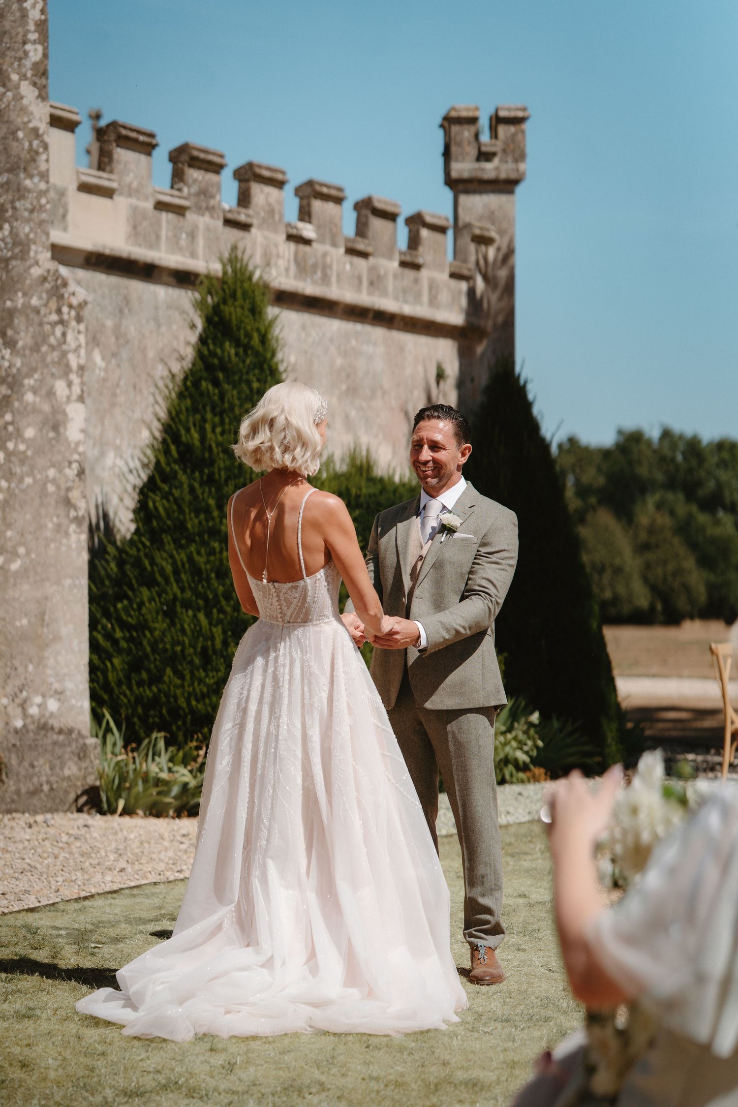 Bride and groom holding hands during outdoor wedding ceremony at a historic castle on a sunny day.