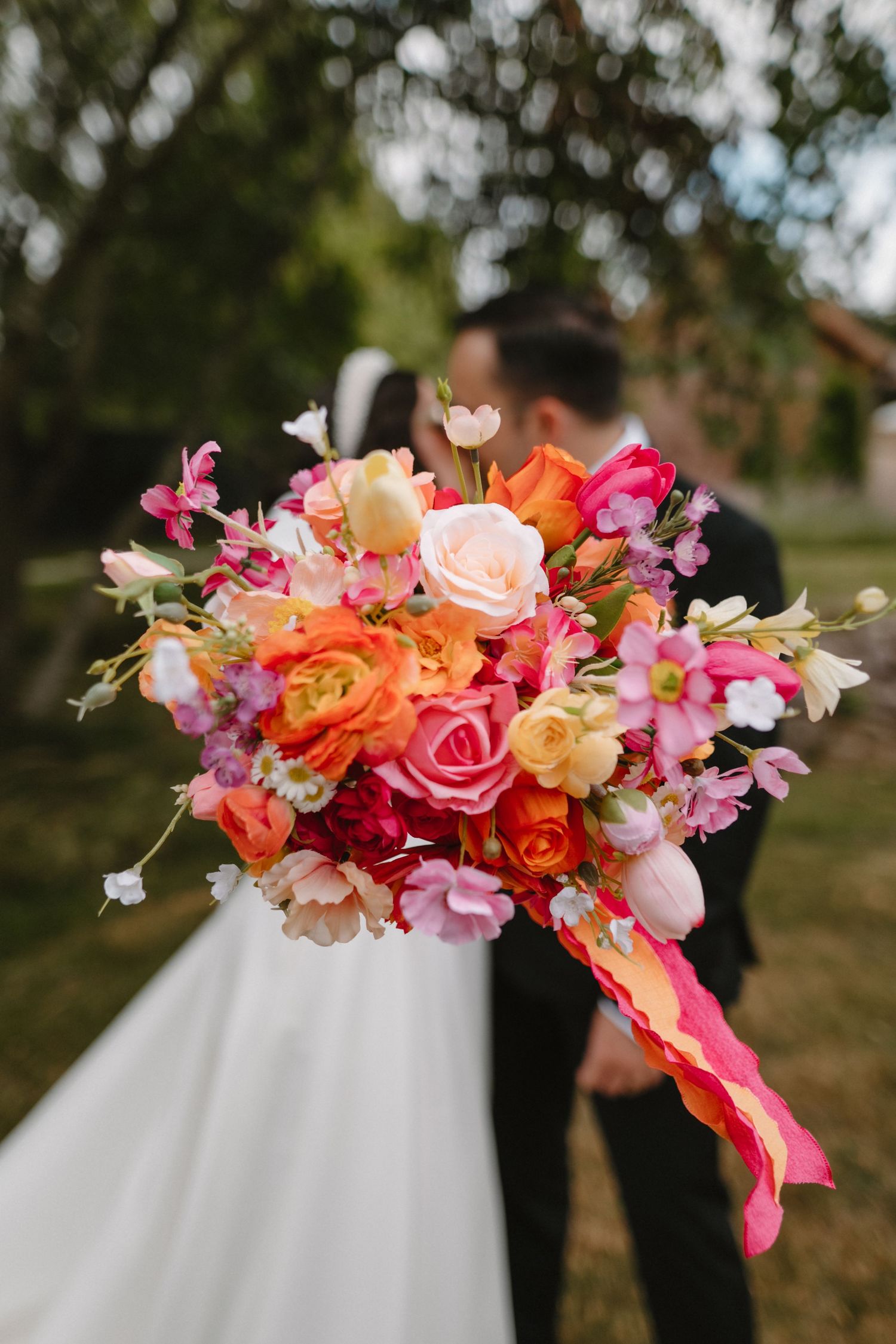 Bride and groom kissing behind a vibrant wedding bouquet of pink, orange, and peach roses outdoors.