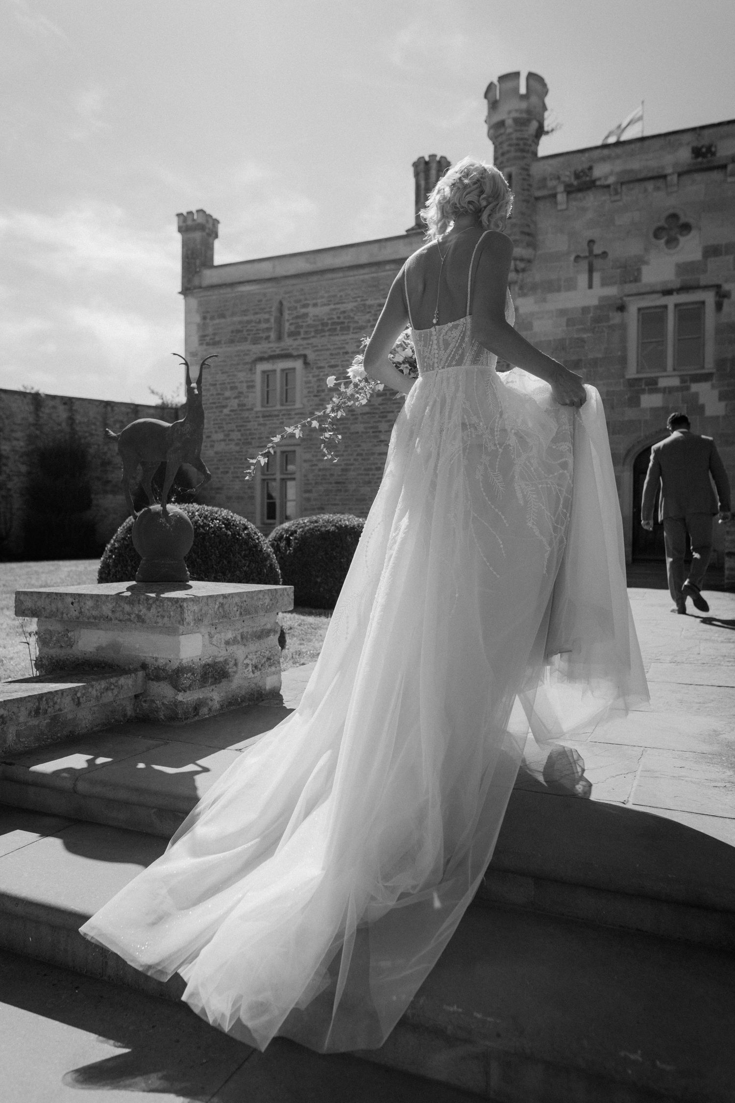 Bride in elegant lace wedding gown with flowing train stands on stone steps outside a historic castle.
