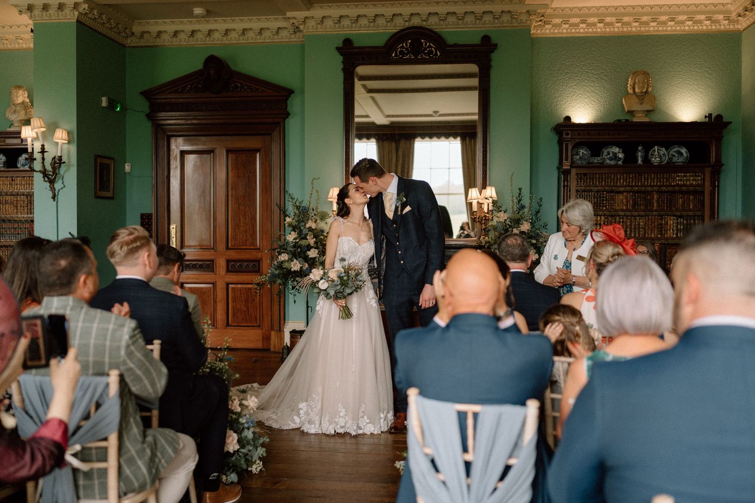 Wedding ceremony in progress in an elegant green room with wooden details and seated guests at Prestwold Hall.