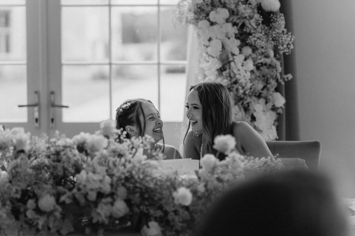 A black and white photograph of a bride captured through flowers at a wedding reception.