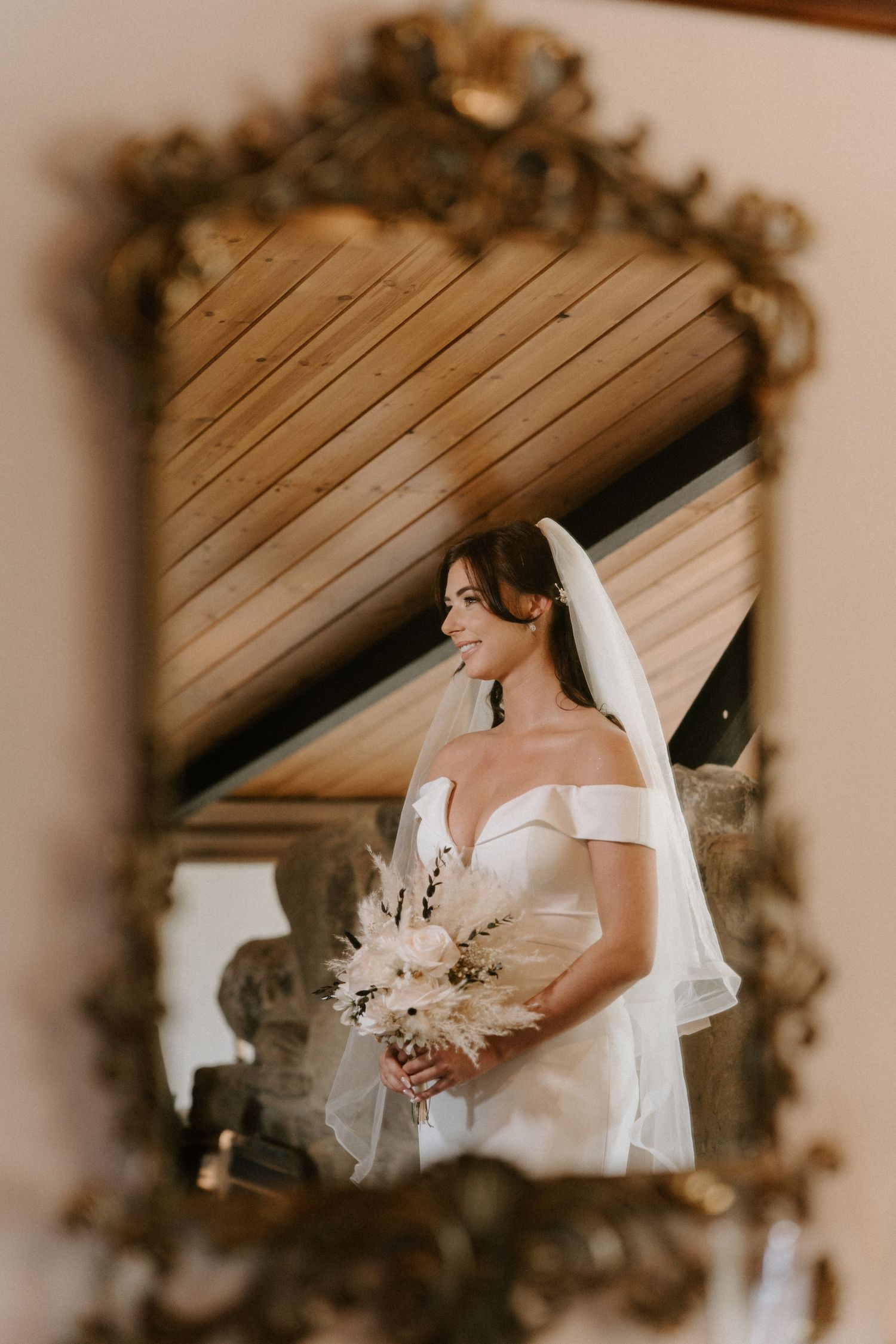 Elegant bridal portrait reflected in an ornate vintage gold mirror showing a white off-shoulder wedding dress and veil.