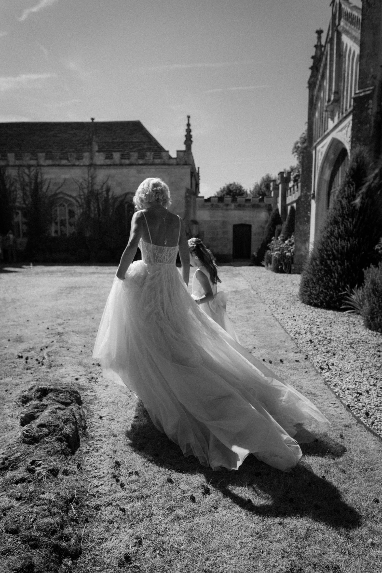 Bride in flowing backless tulle wedding gown walks with groom across grounds of grand English estate.