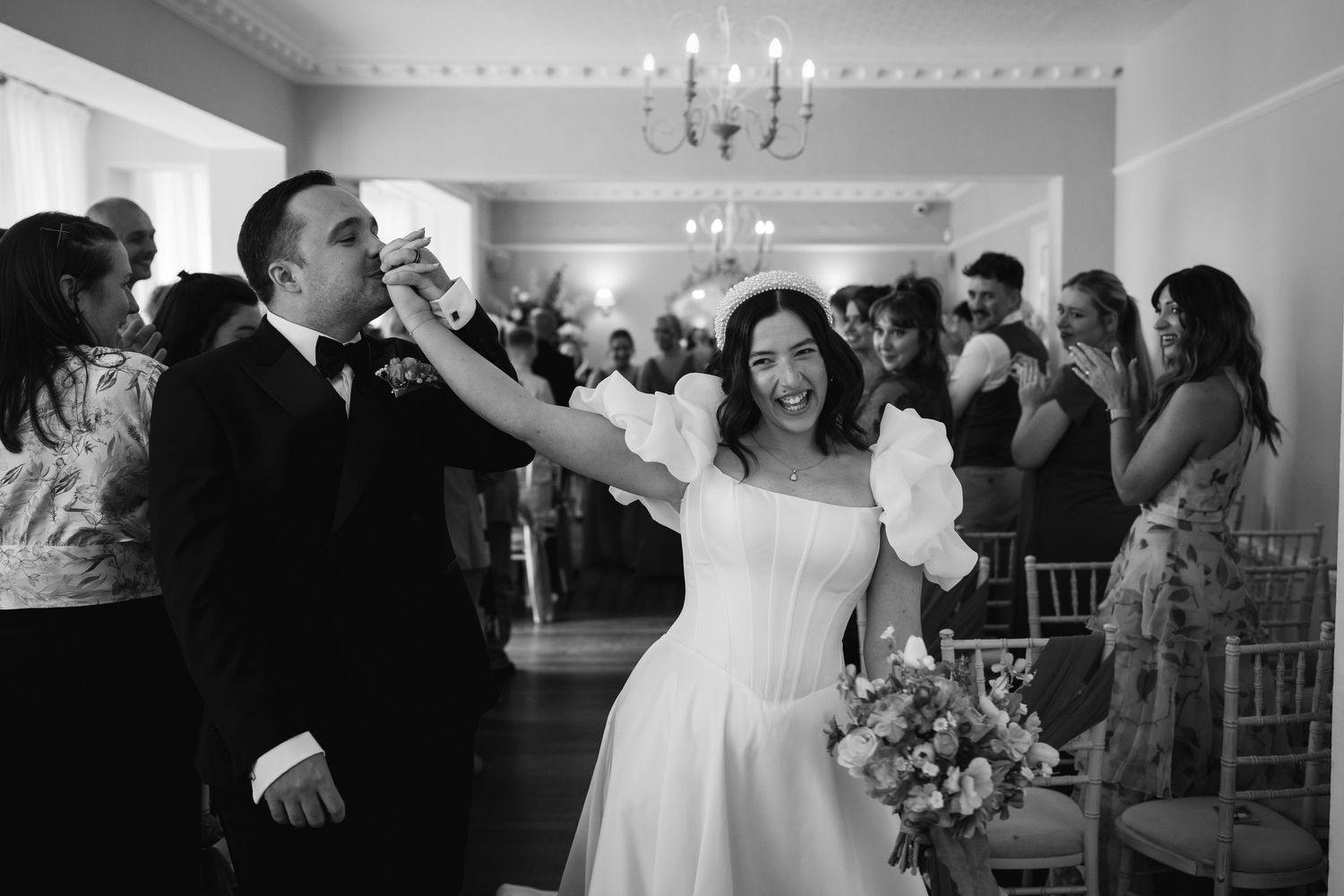Black and white photo of a bride and groom celebrating their wedding reception, bride holding bouquet while guests cheer.