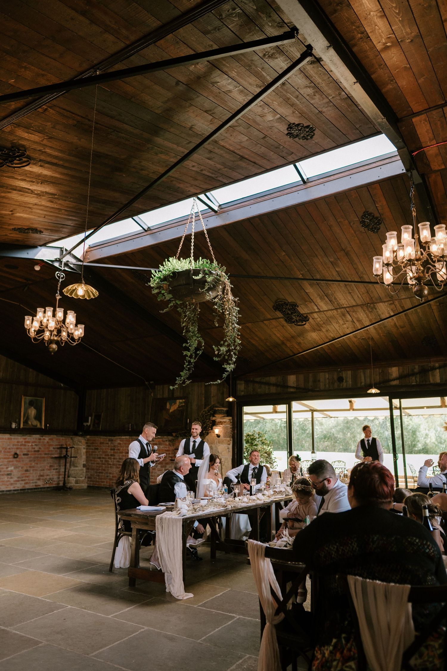A rustic wedding reception venue with wooden beam ceiling, chandeliers, skylights and hanging plants in an elegant setting.
