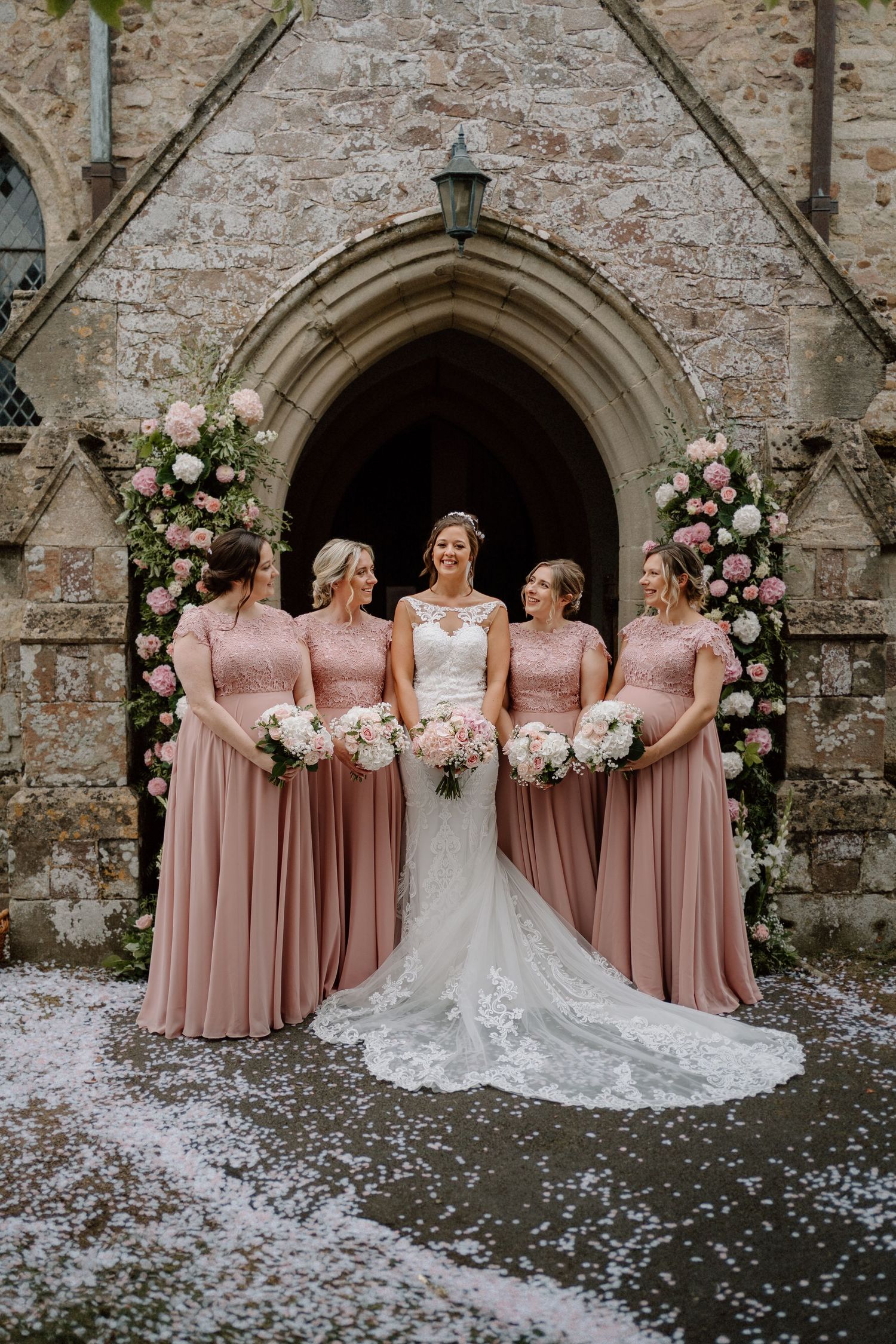 Bride and bridesmaids in dusty pink dresses pose under stone church archway decorated with pink and white floral arrangements.