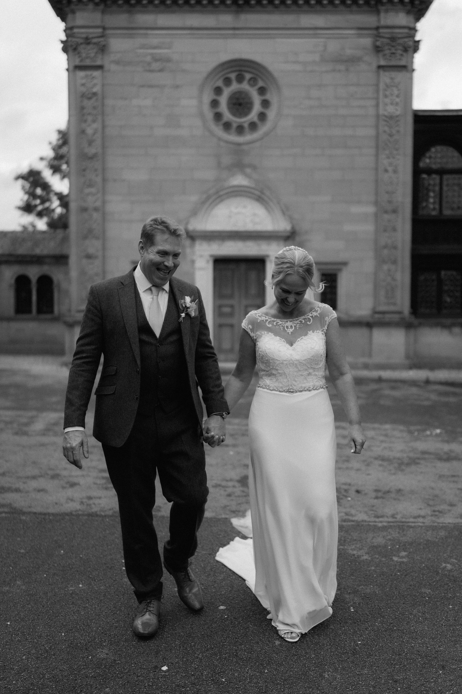 A wedding couple walking together outside a historic stone church building with arched windows at Bourton Hall.