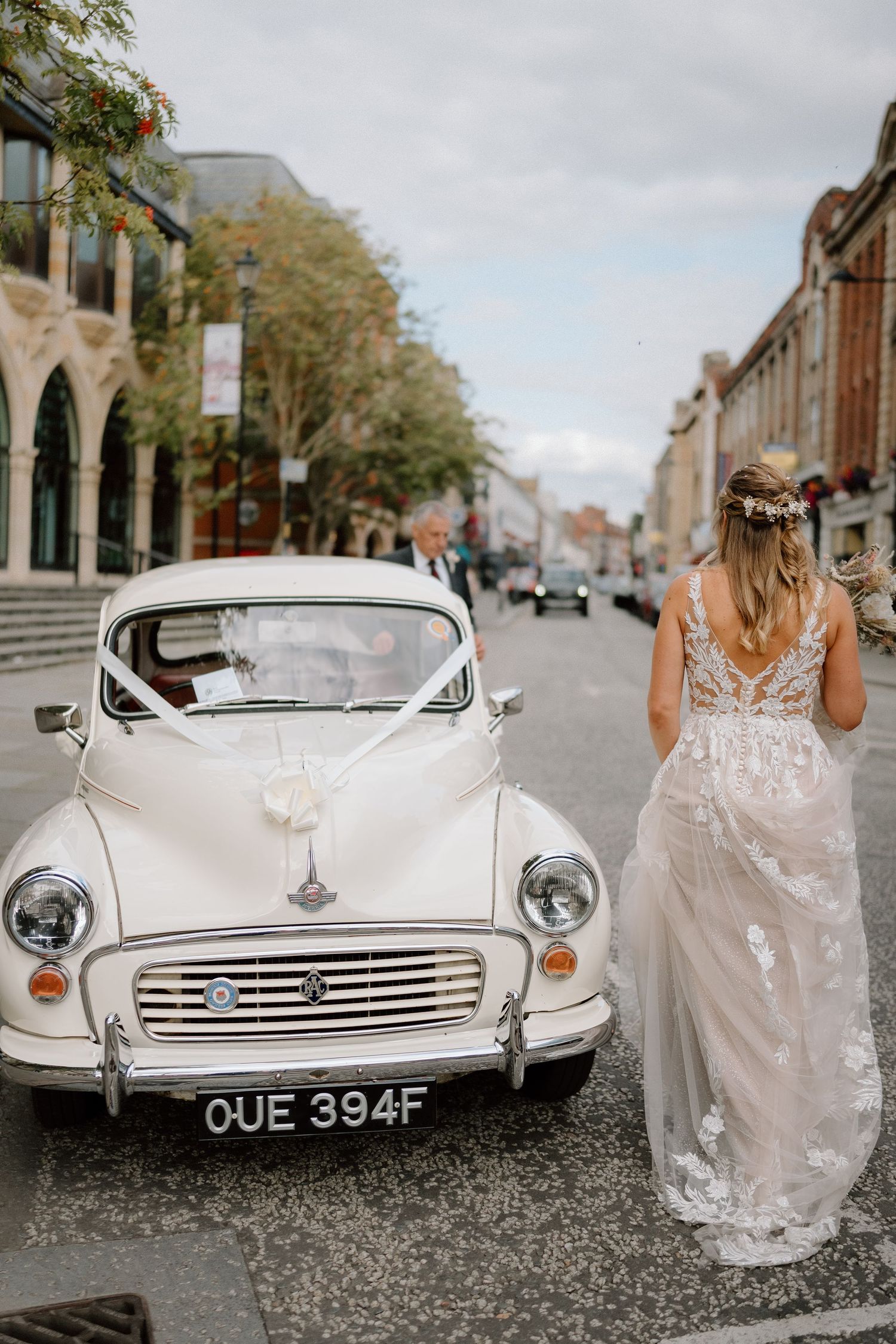 A bride stands beside a vintage white Morris Minor car decorated for a wedding in Northampton.