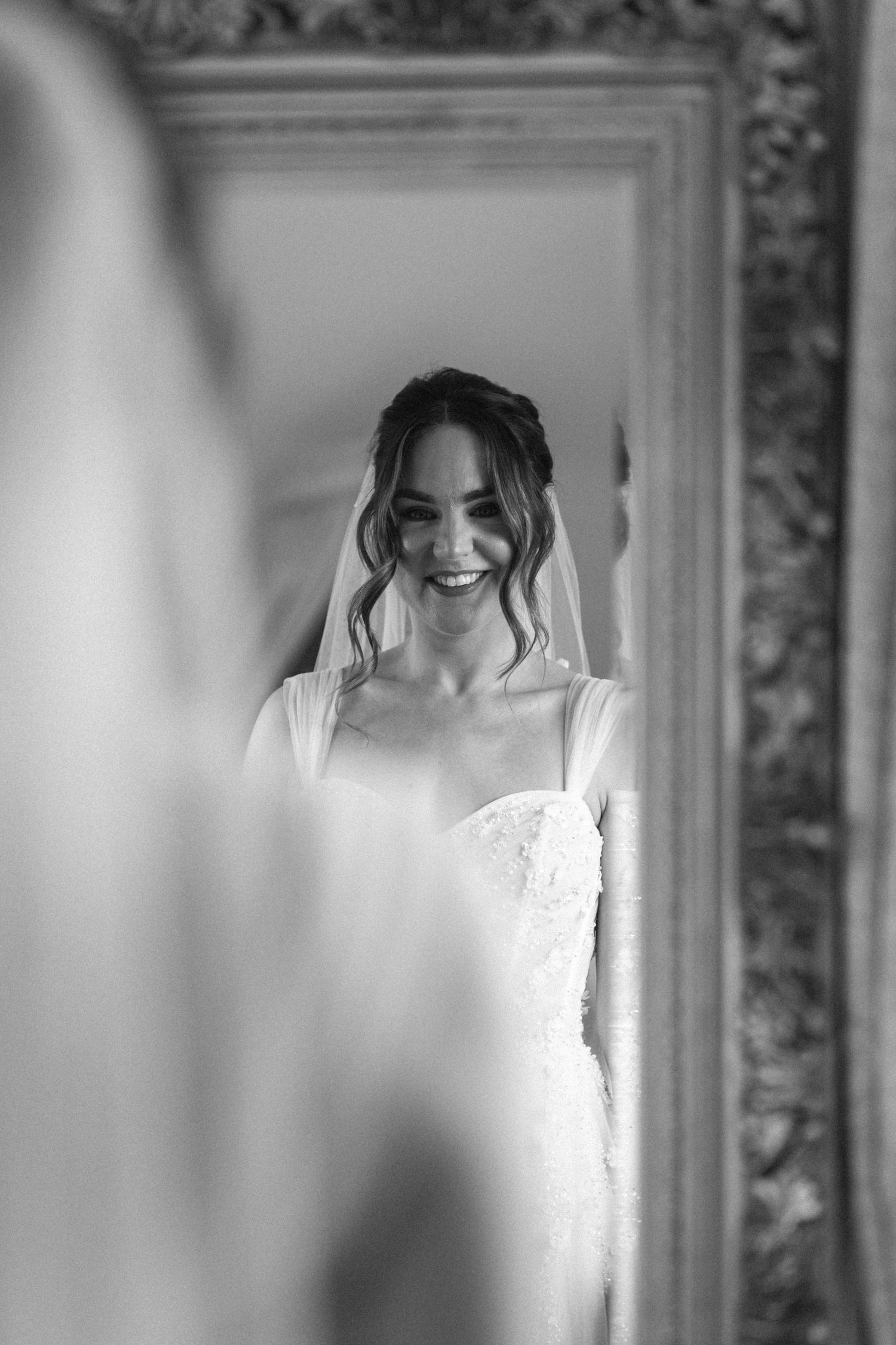 Black and white photo of a smiling bride in a lace gown and veil, reflected in an ornate gold-framed mirror.