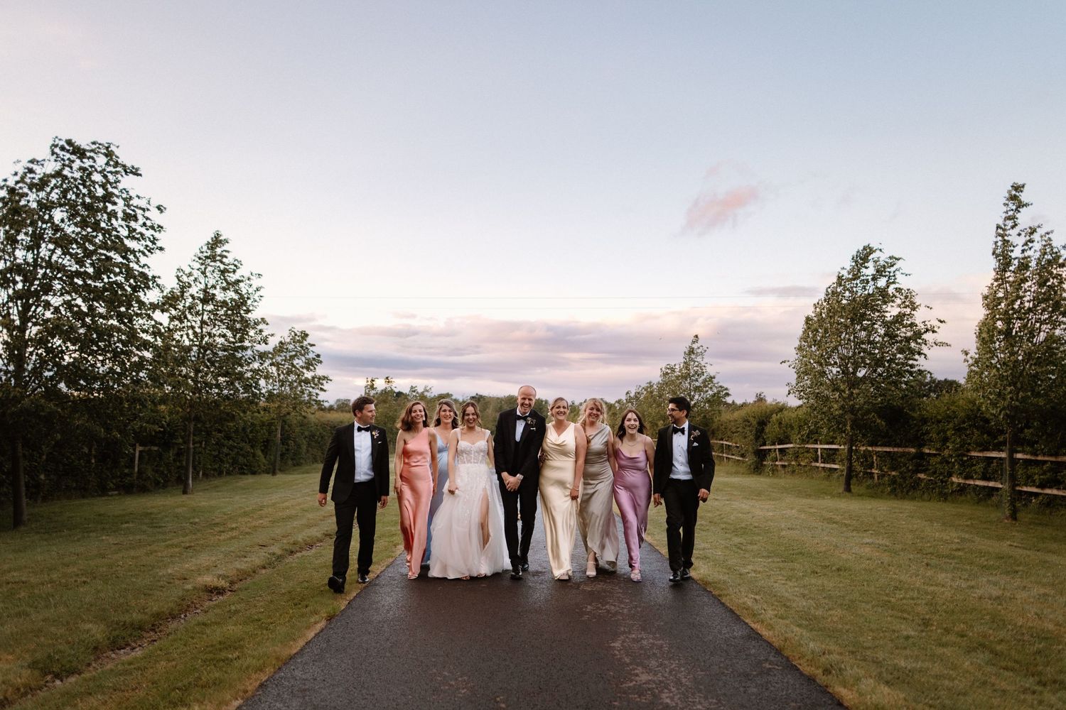 Wedding party walks down a tree-lined path at sunset, bride in white gown with bridesmaids in pink and purple dresses.