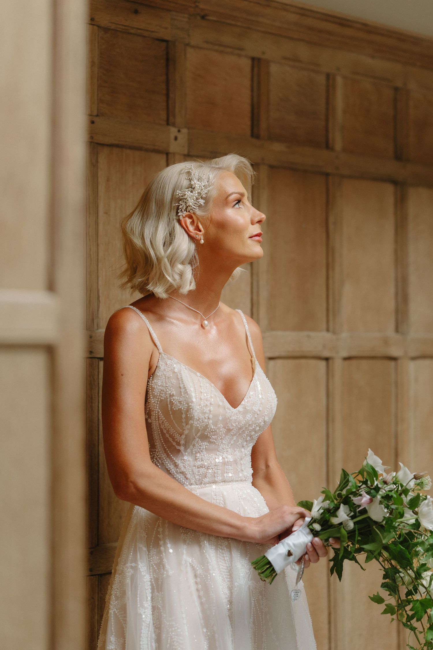 Elegant bride in beaded gown holding bouquet, blonde hair with floral accessory, gazing upward in a wood-paneled room.