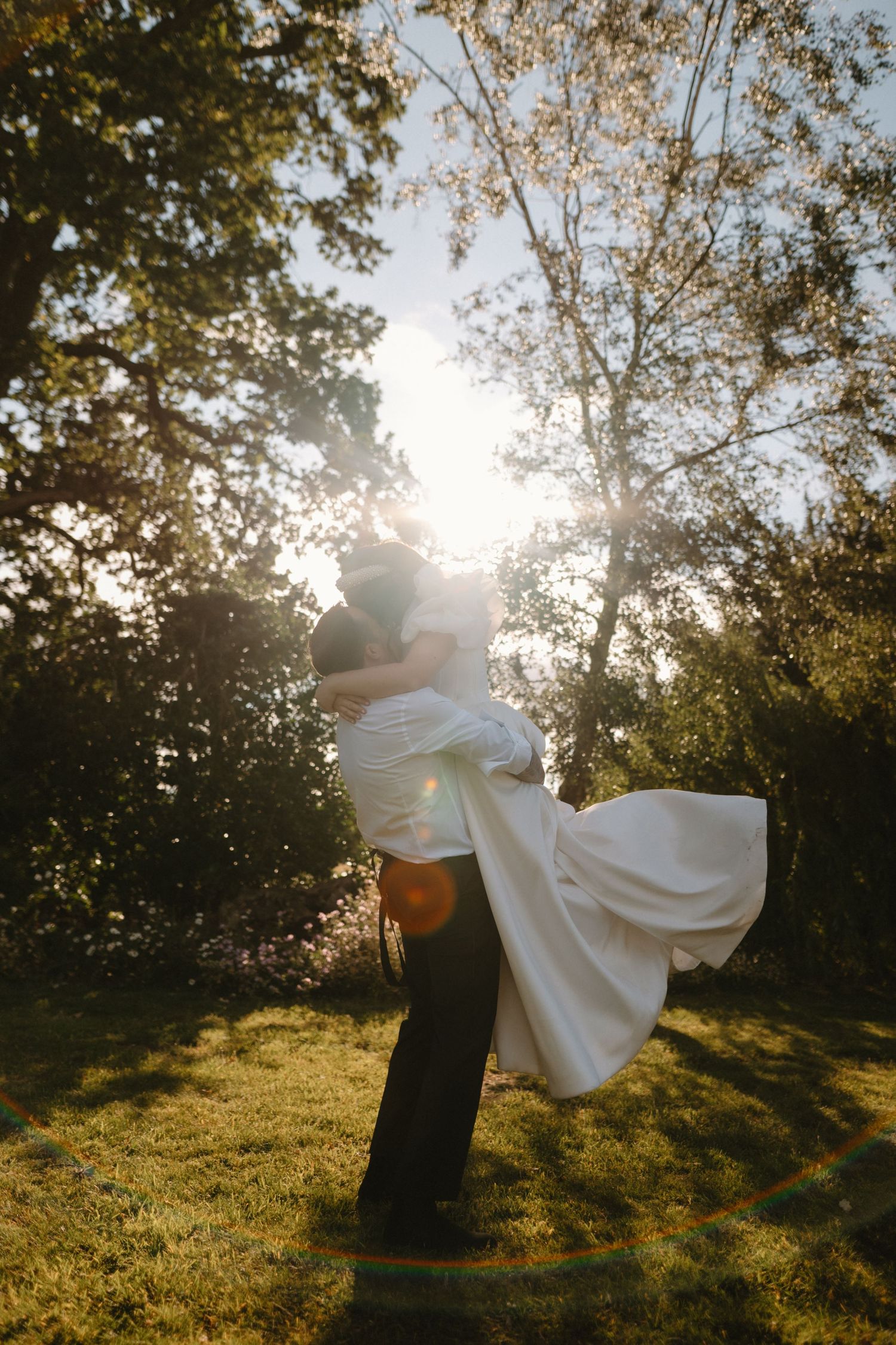 Groom lifting bride in white gown outdoors, sun flare through trees, romantic golden hour wedding photo.