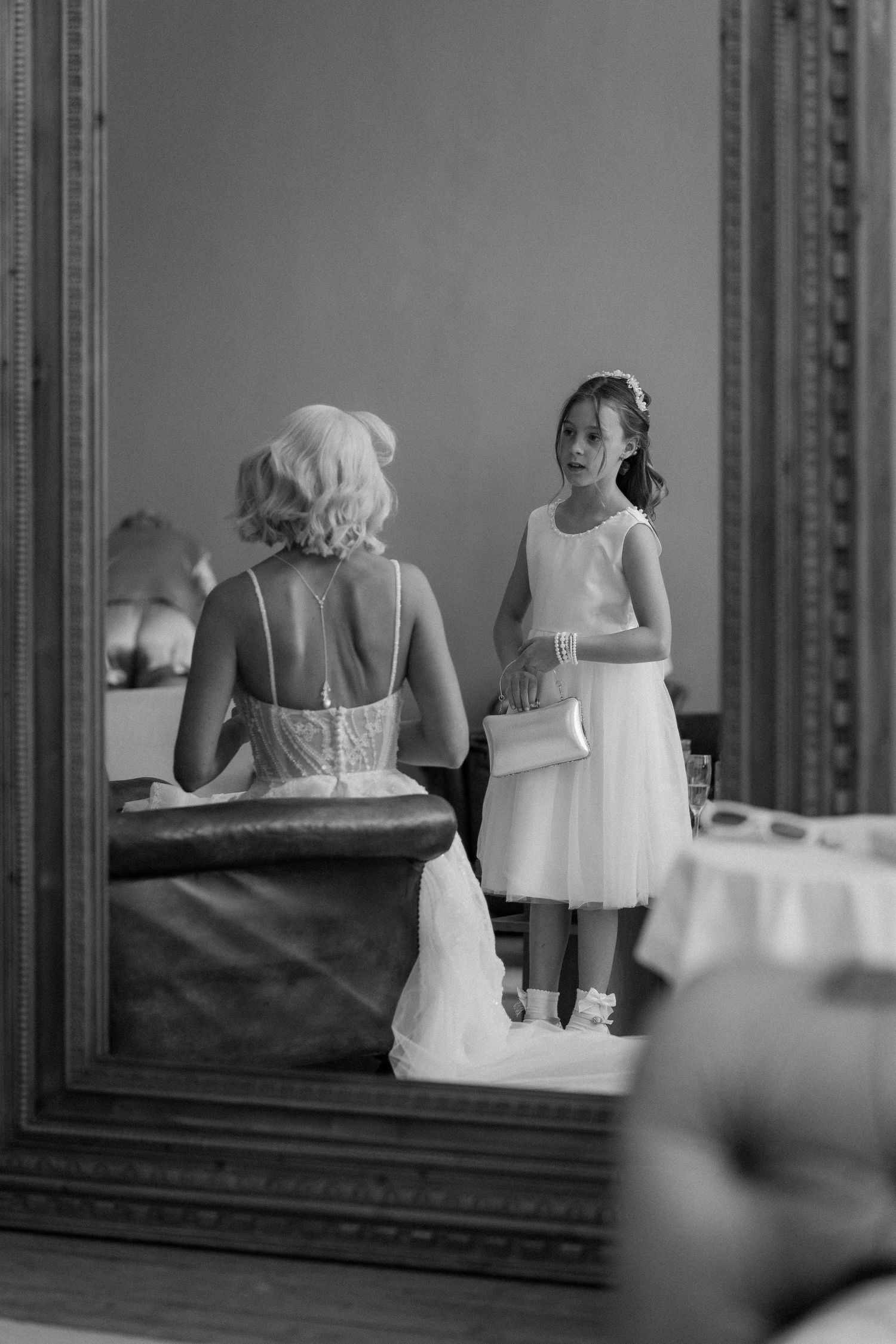 Black and white photo of a flower girl in white dress holding purse, reflected in ornate mirror beside bride getting ready.