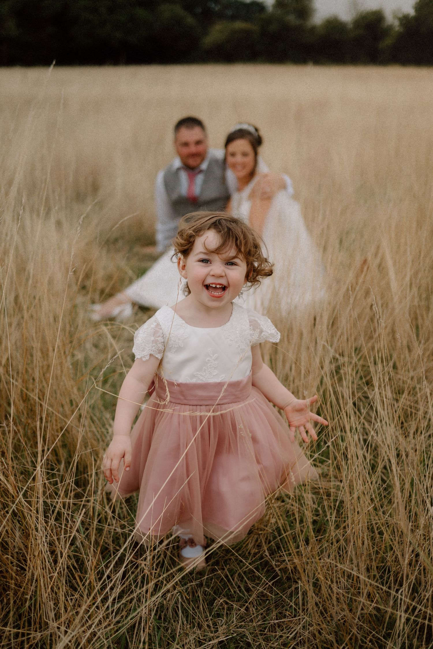 A joyful child in a tulle dress runs through a golden wheat field while a wedding couple looks on in the background.