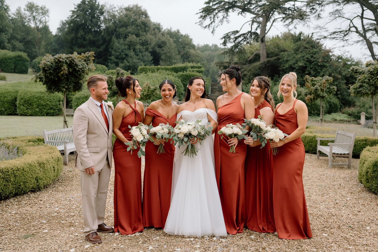 A wedding party poses outdoors with bridesmaids in burnt orange dresses on a gravel path at Harlaxton Manor.