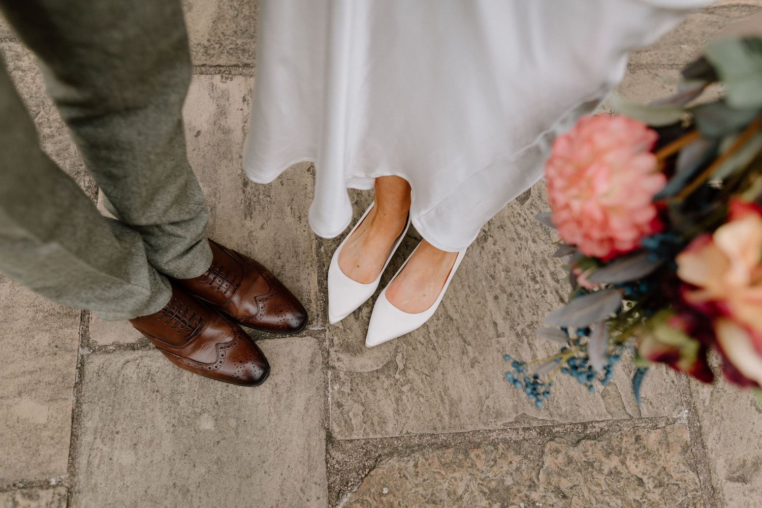 Brown dress shoes and beige flats stand next to pink and coral flowers on stone ground in Coventry.